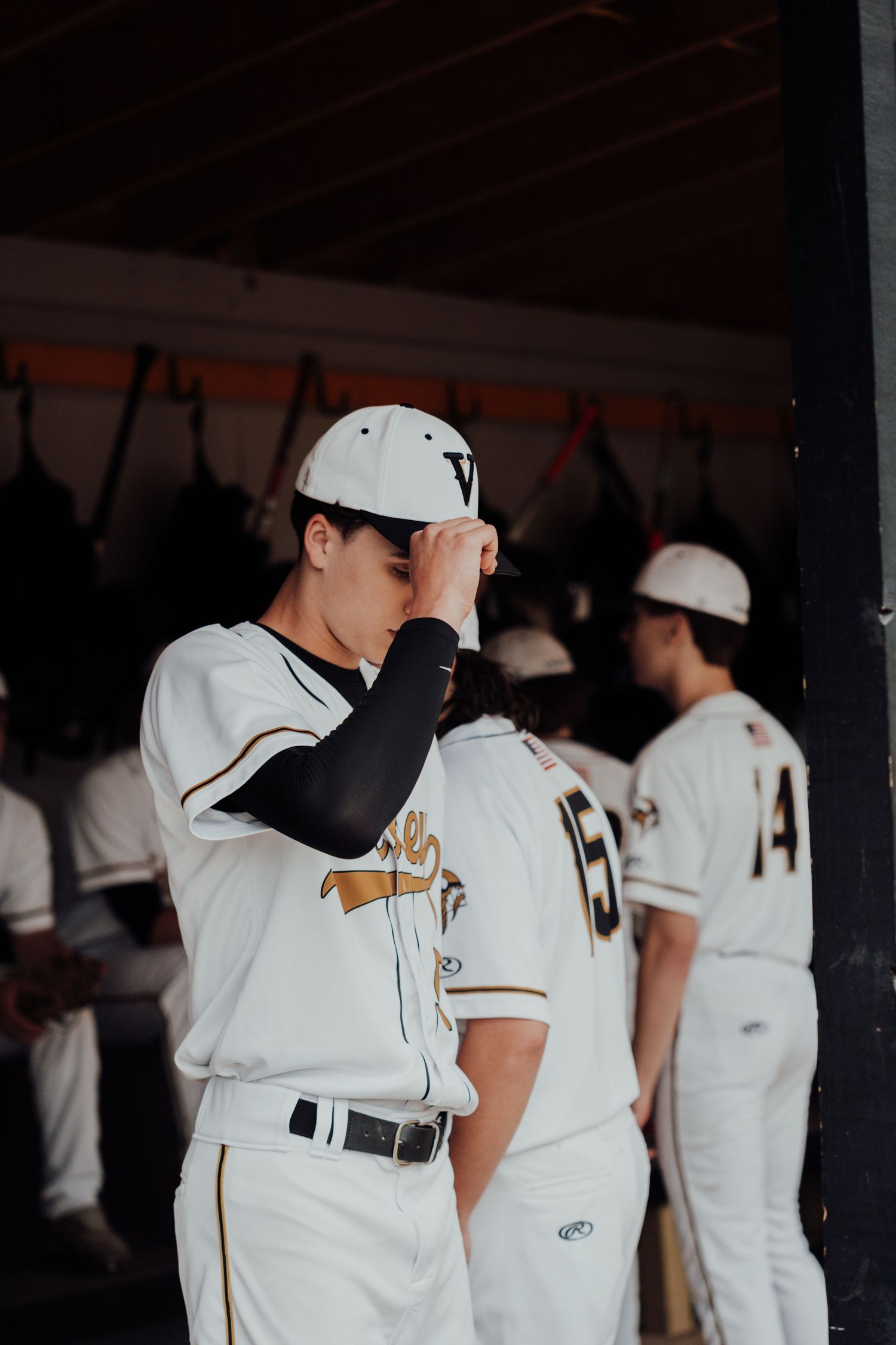 Baseball players in white uniforms stand together in shaded dugout area.