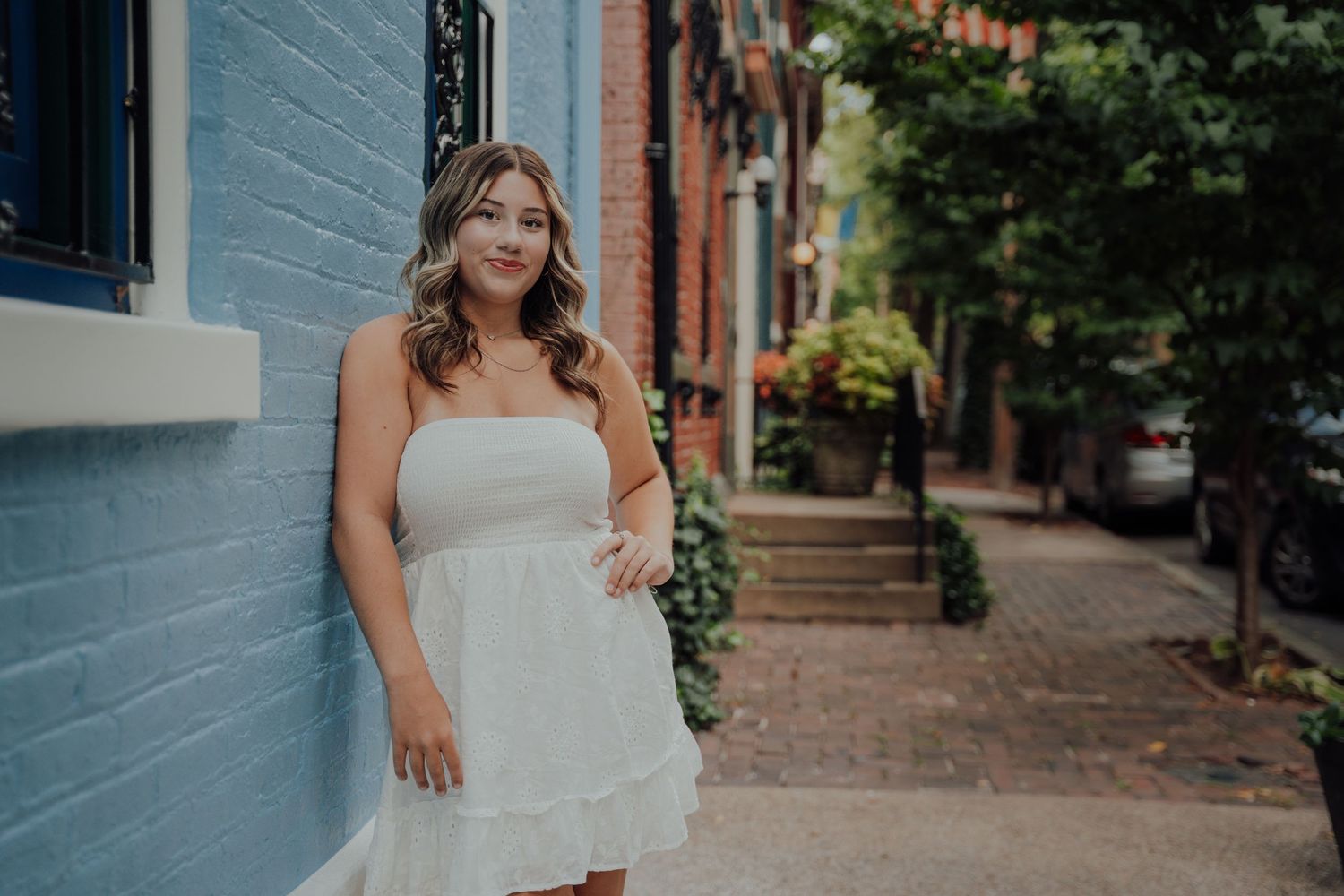 A person in a white strapless dress stands smiling on a charming brick-lined residential street with blue buildings.