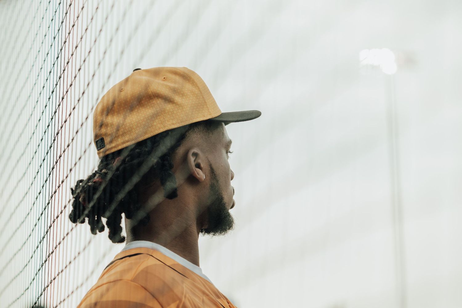 Side profile of someone wearing a tan cap and orange shirt gazing upward against a light background.
