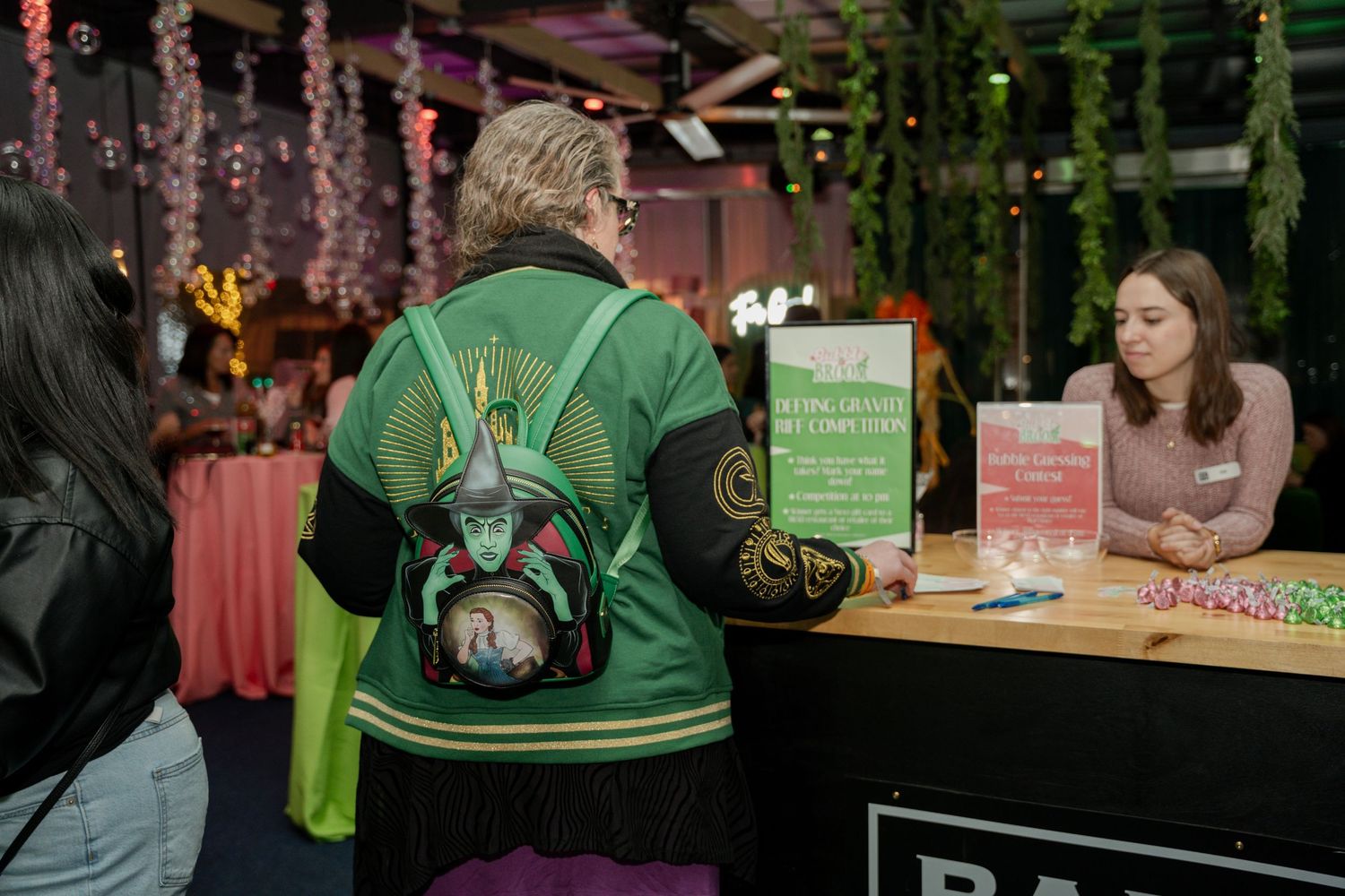Person wearing a bright green backpack stands at a book signing event with pink decor and displays.