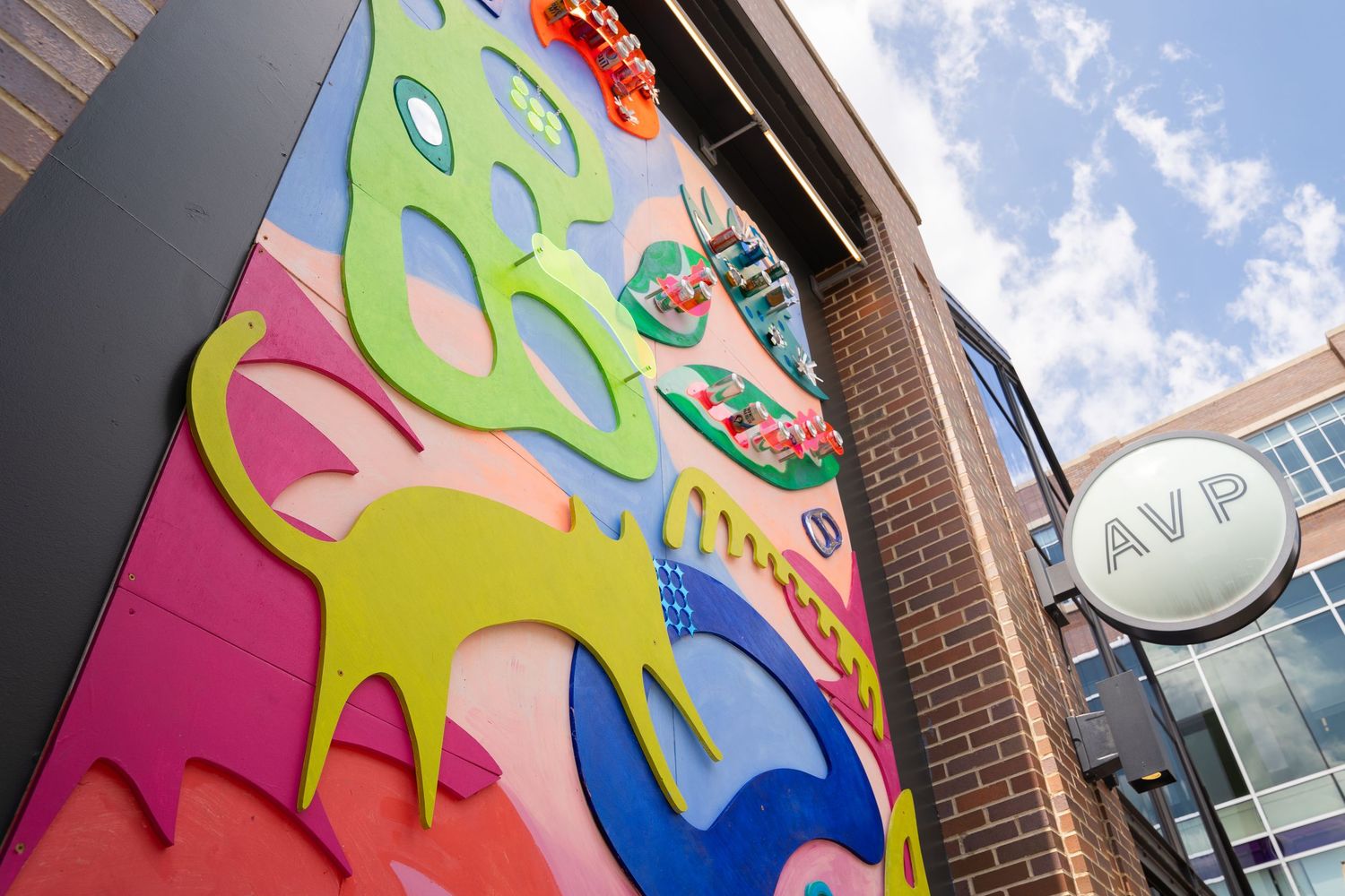 Colorful street art mural with abstract shapes and bright patterns adorns the side of a brick building under blue sky.