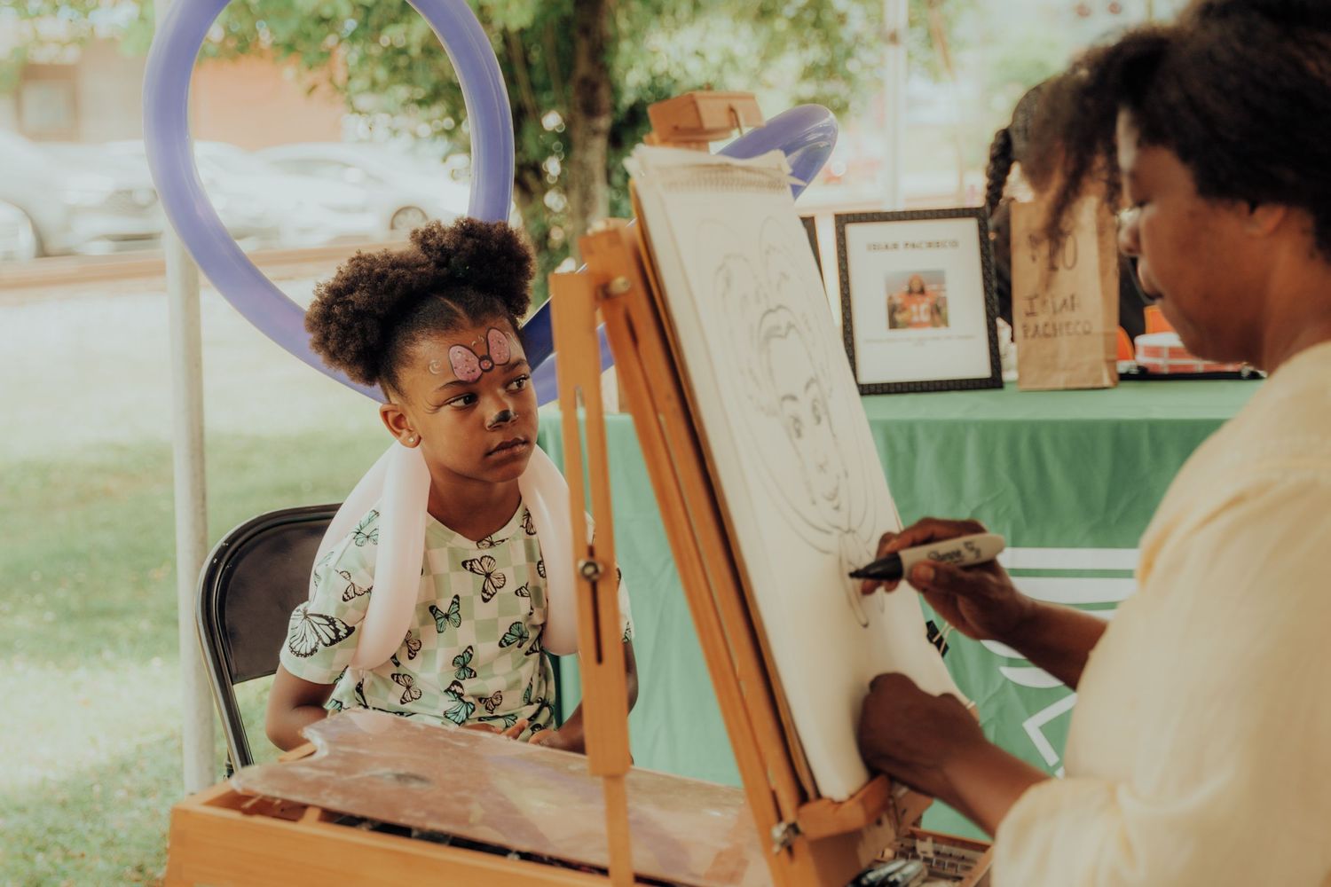 Artist sketches on an easel while sitting across from someone during an outdoor portrait session in a garden setting.