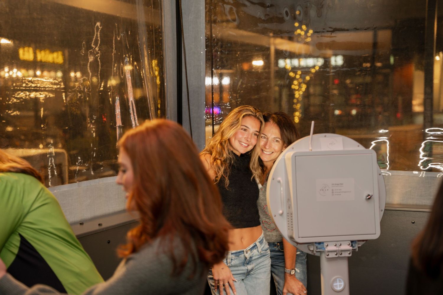 Party guests gather around camera equipment in a dimly lit venue with neon lights visible through windows.