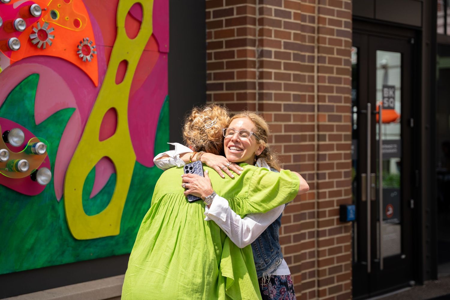 Two people share a warm embrace in front of a colorful mural on a brick wall outside a building.