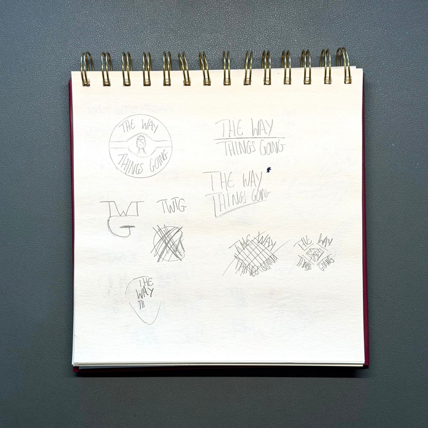 A spiral-bound notebook with hand-drawn logo sketches and doodles on white paper against a dark background.
