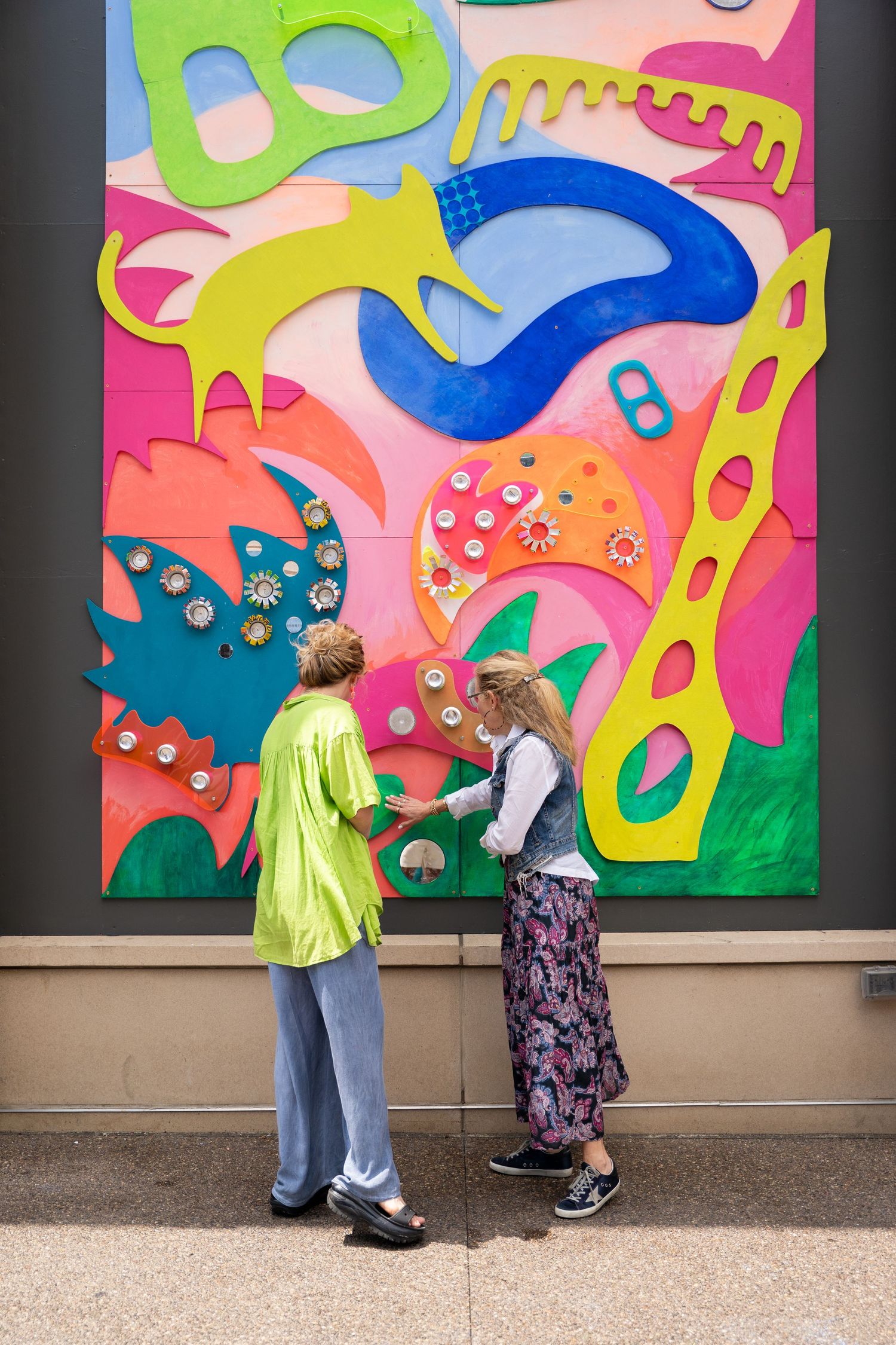 Two people observe a vibrant abstract mural featuring colorful organic shapes and patterns on a wall.
