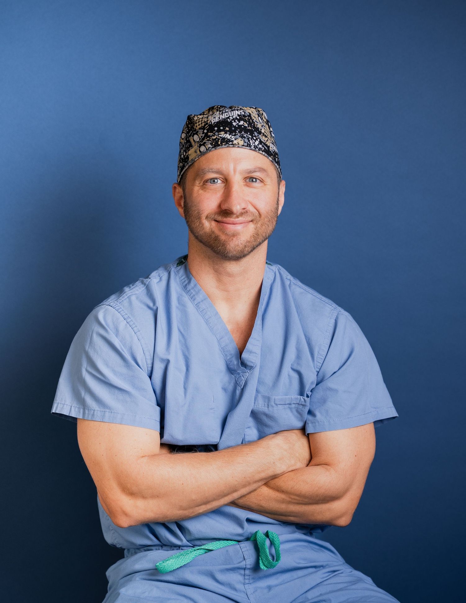Healthcare professional in light blue scrubs and patterned cap poses with crossed arms against blue background.