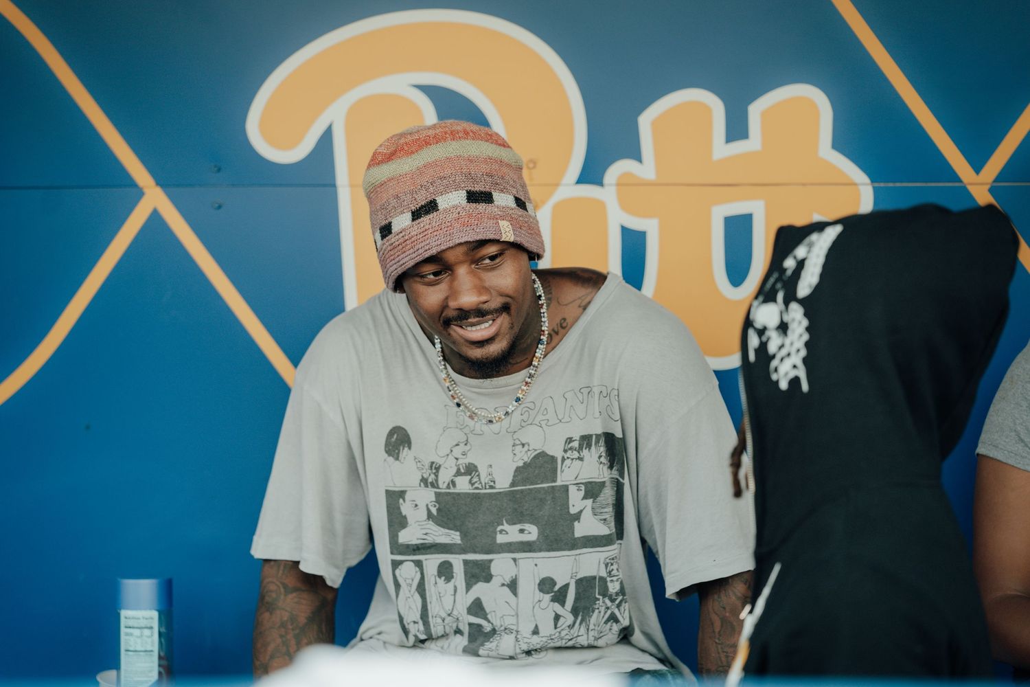A person wearing a beanie and graphic t-shirt smiles joyfully while sitting in front of a blue wall with geometric patterns.