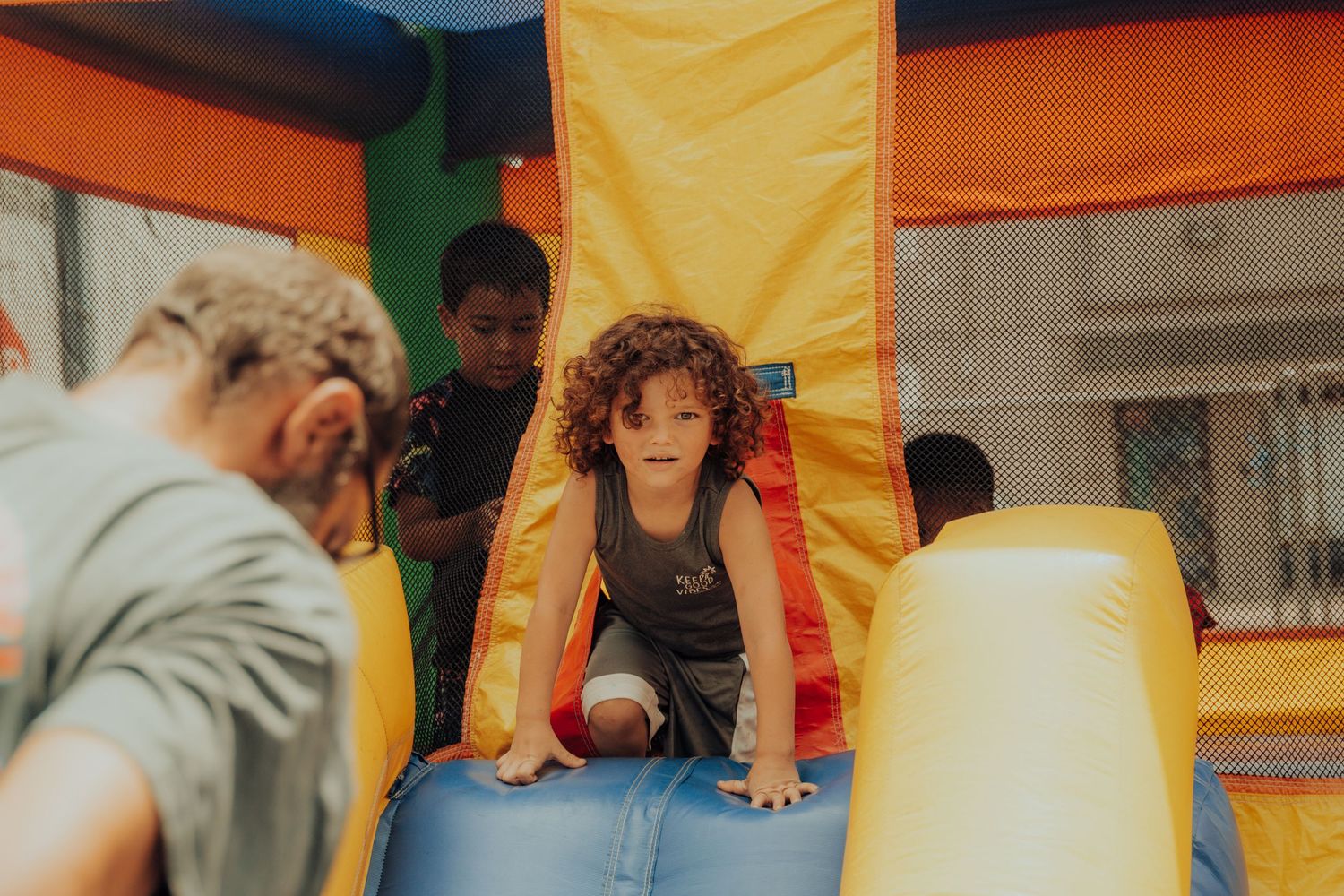 A child climbs through an inflatable bounce house slide with yellow and orange details as others look on.
