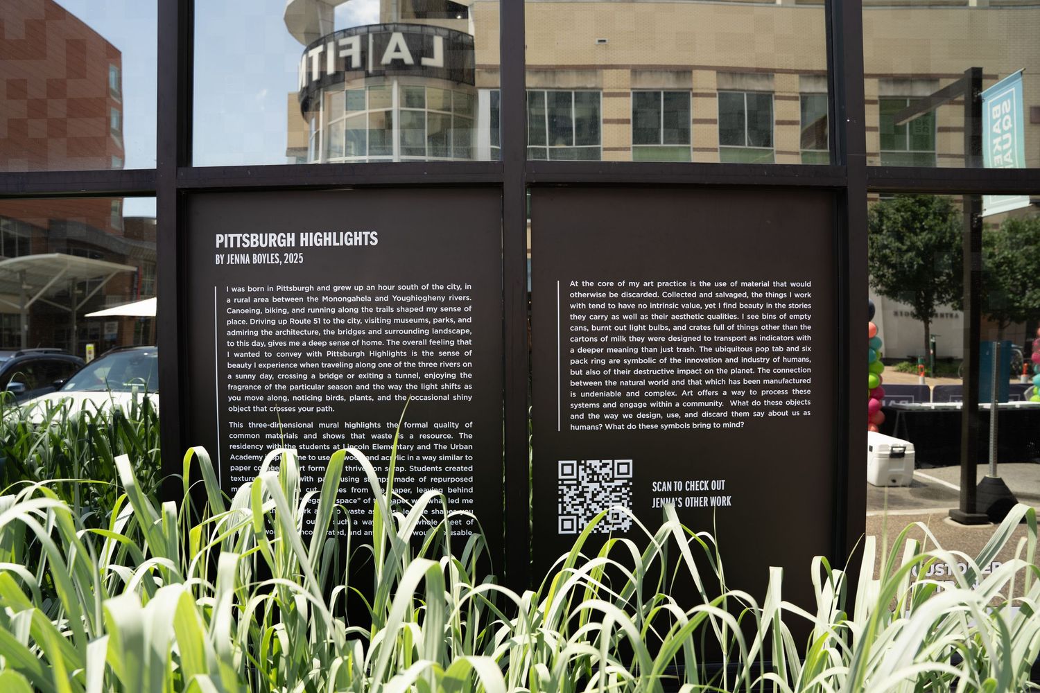 Black interpretive signage stands in a garden bed with tall ornamental grasses near a city building.