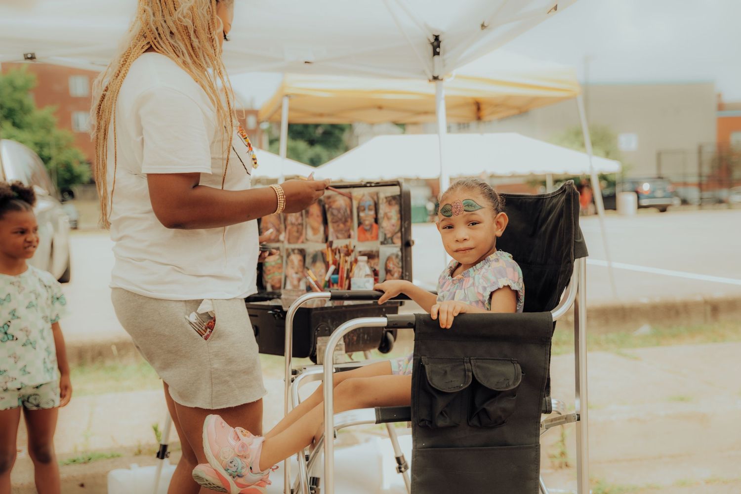 A child sits in a portable camping chair at an outdoor event on a sunny day.