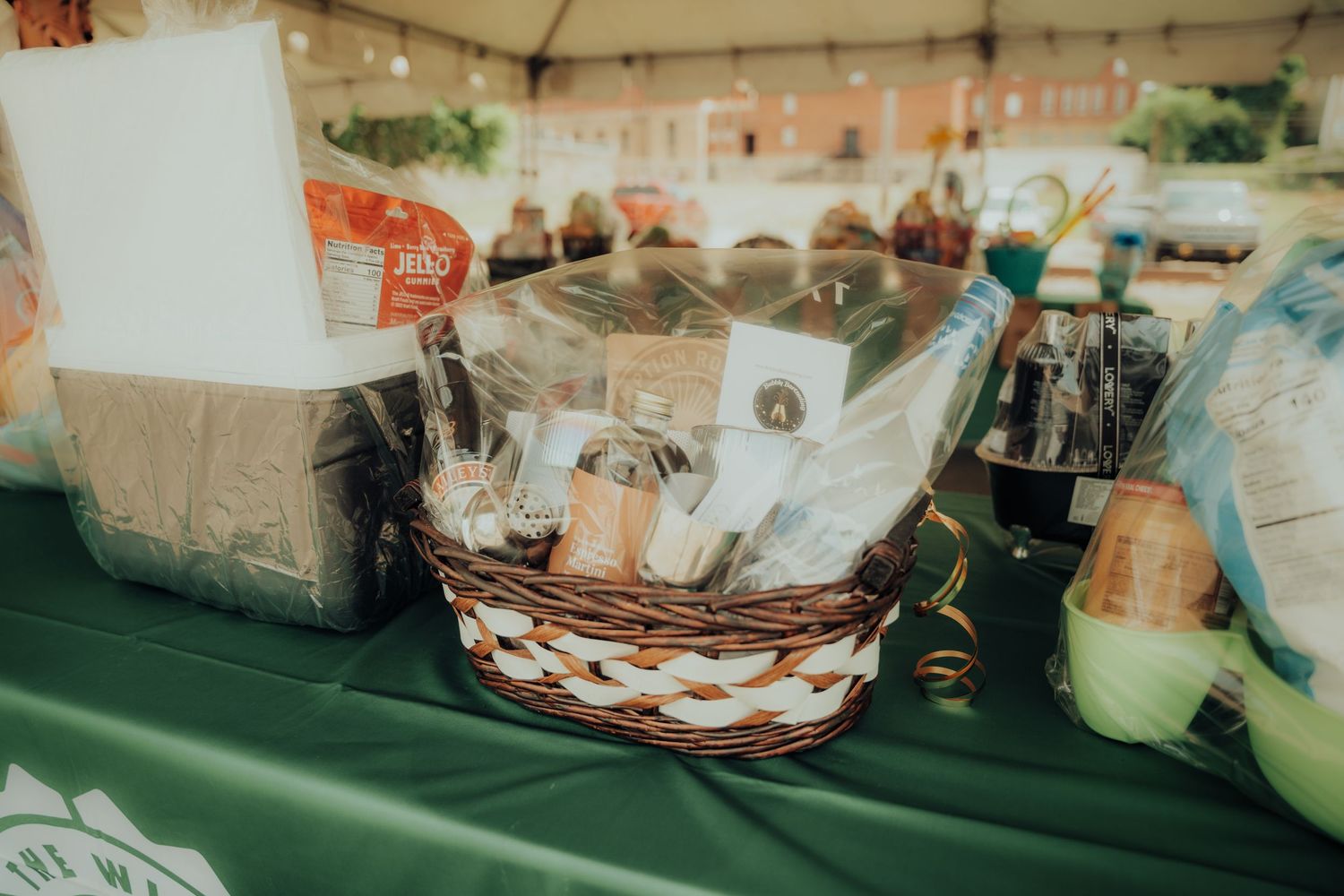 Assorted condiments and supplies organized in a woven basket on a green tablecloth at an outdoor event.