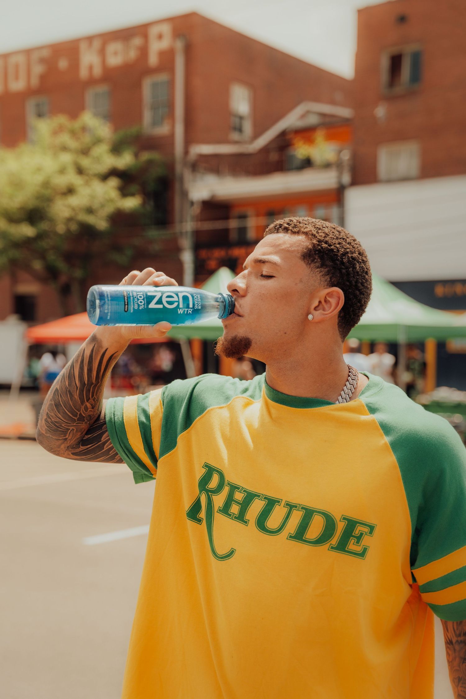 Someone wearing a yellow and green Rhude sweatshirt drinks from a blue Zen beverage can in an urban setting.