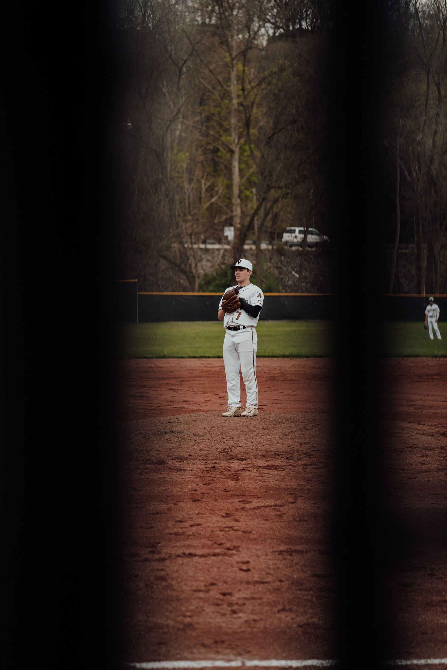 Baseball player in white uniform stands on dirt infield during game, viewed through fence at baseball diamond.