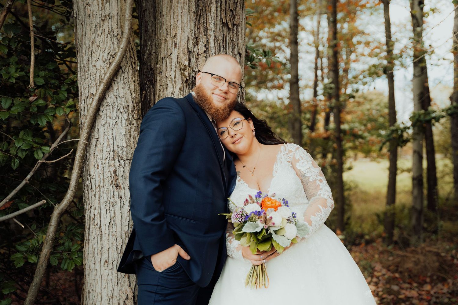 A joyful wedding couple embraces against a tree trunk in an autumn forest setting with a floral bouquet.