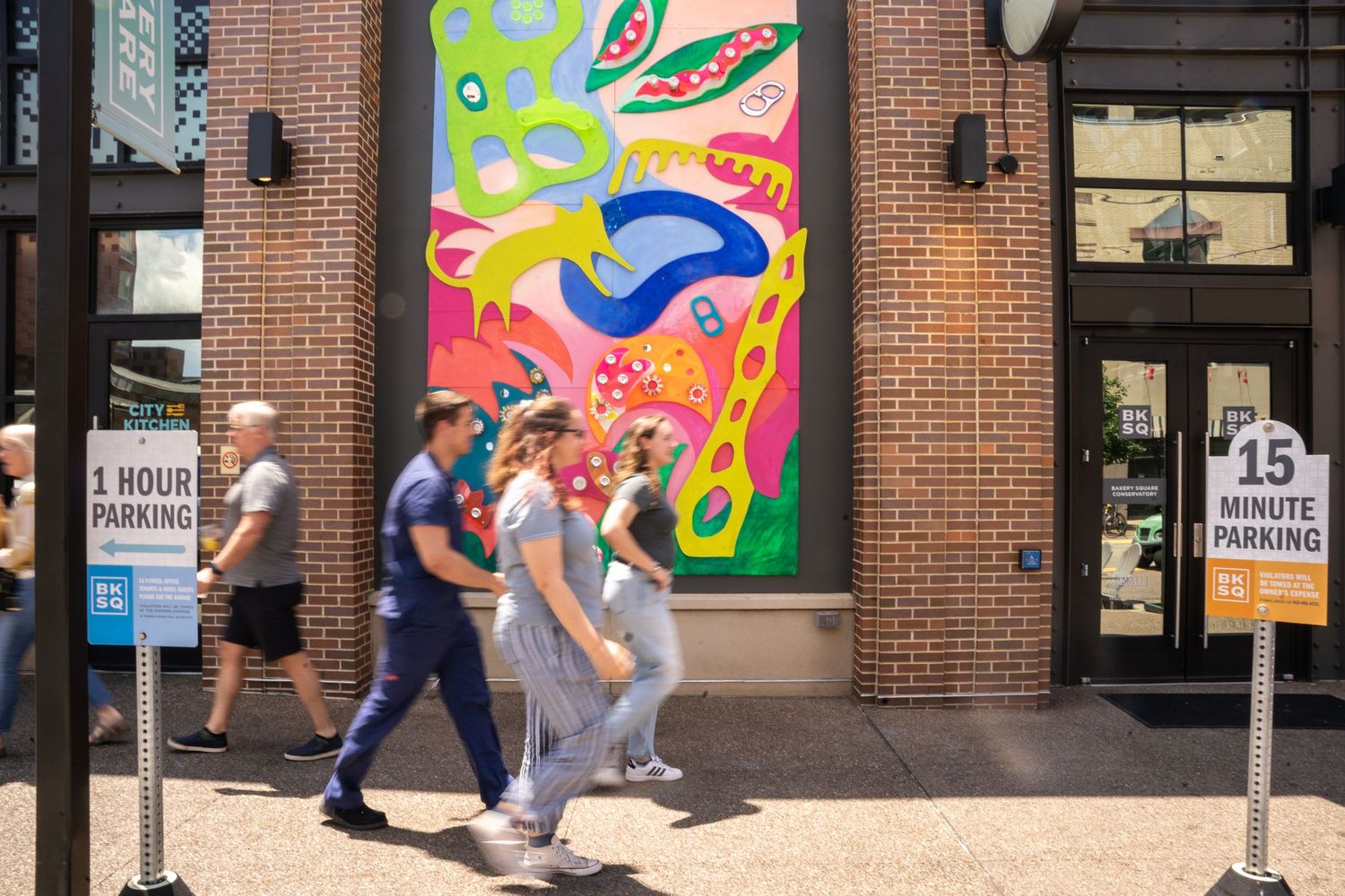 Pedestrians walk past a large colorful abstract mural on a brick wall in an urban setting.