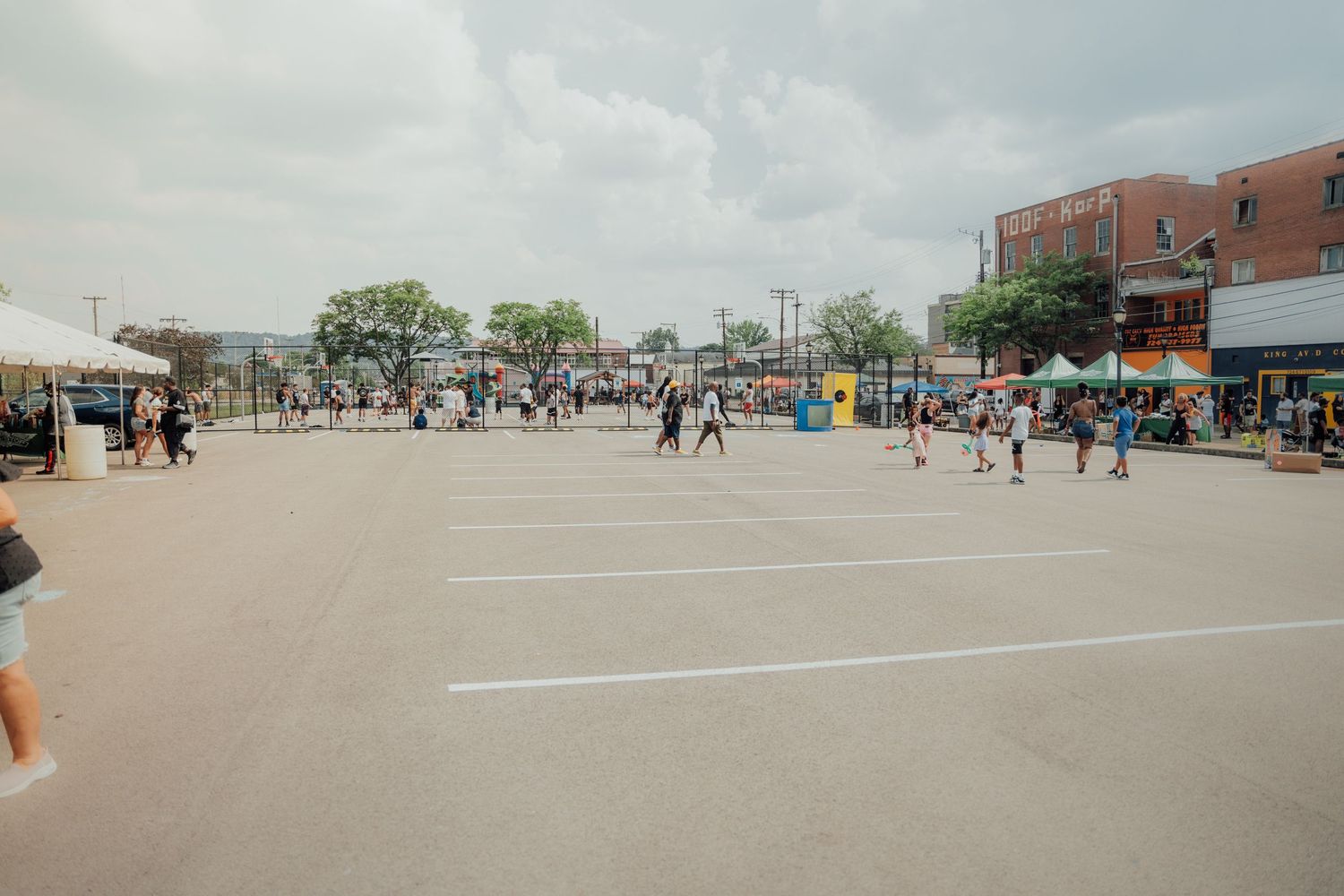 Panoramic view of a shopping center parking lot with scattered cars and pedestrians walking under cloudy skies.
