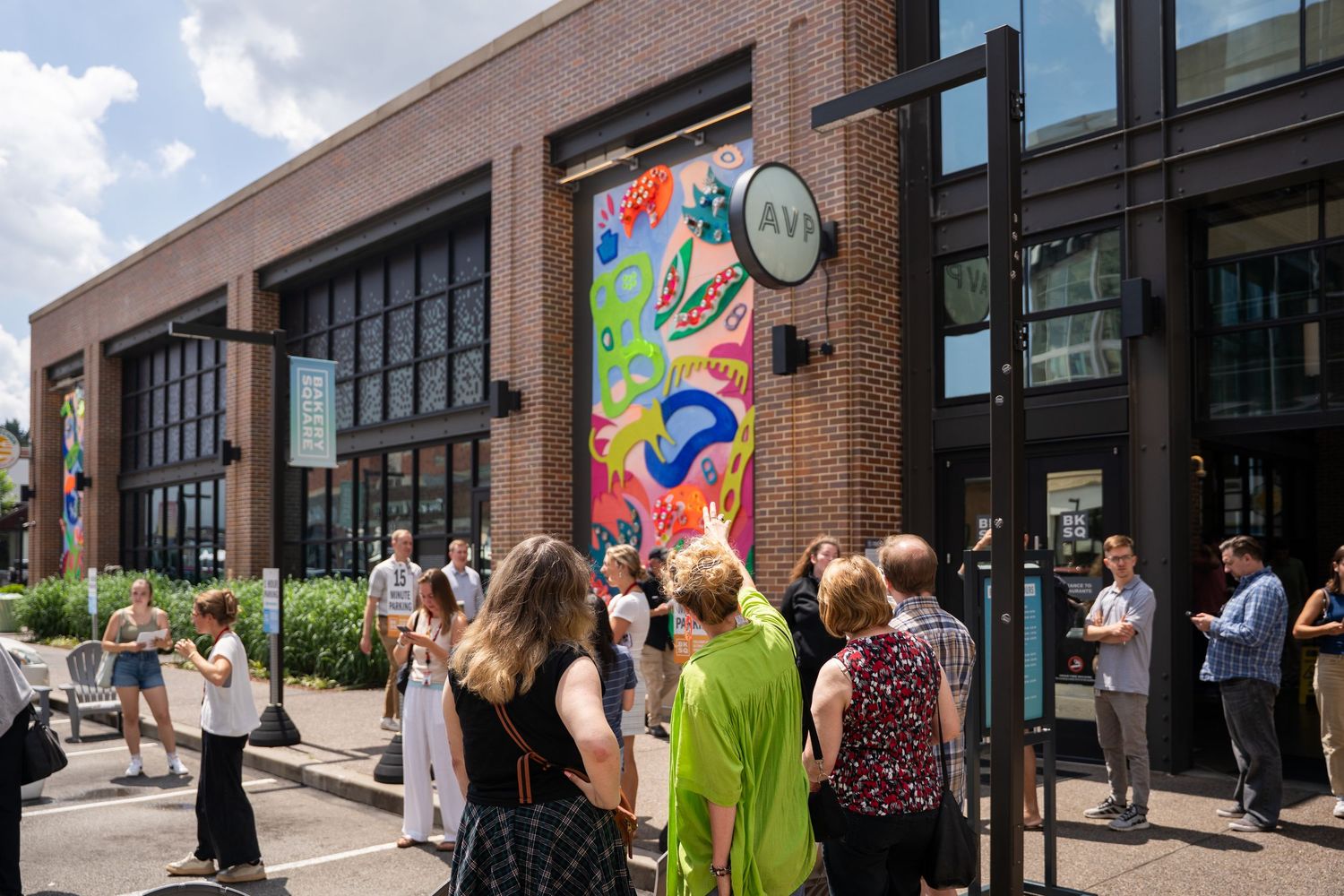 People gather outside a brick building with colorful street art mural during a daytime event.