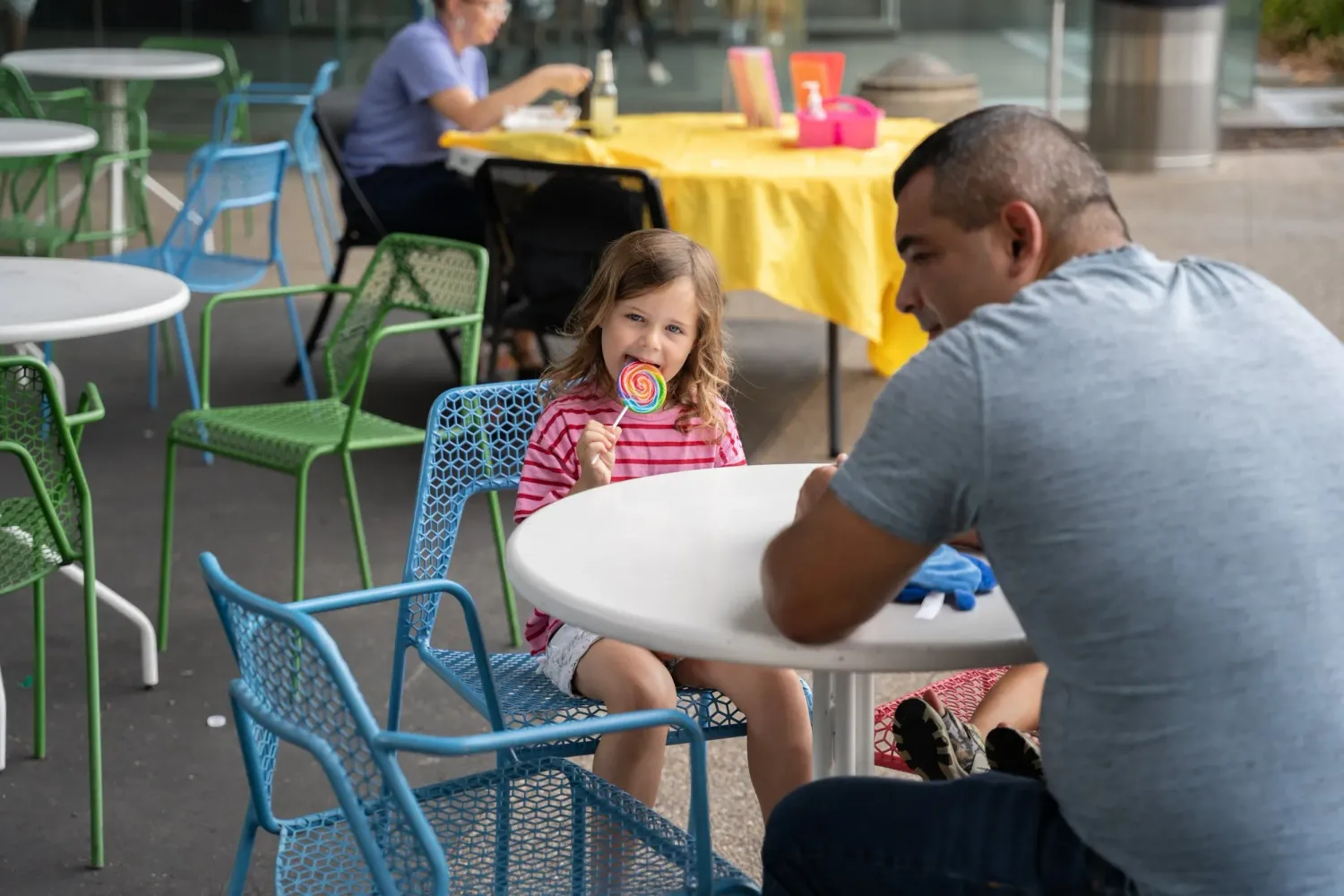 People sit at turquoise metal chairs around tables at an outdoor dining area with colorful yellow tablecloths.