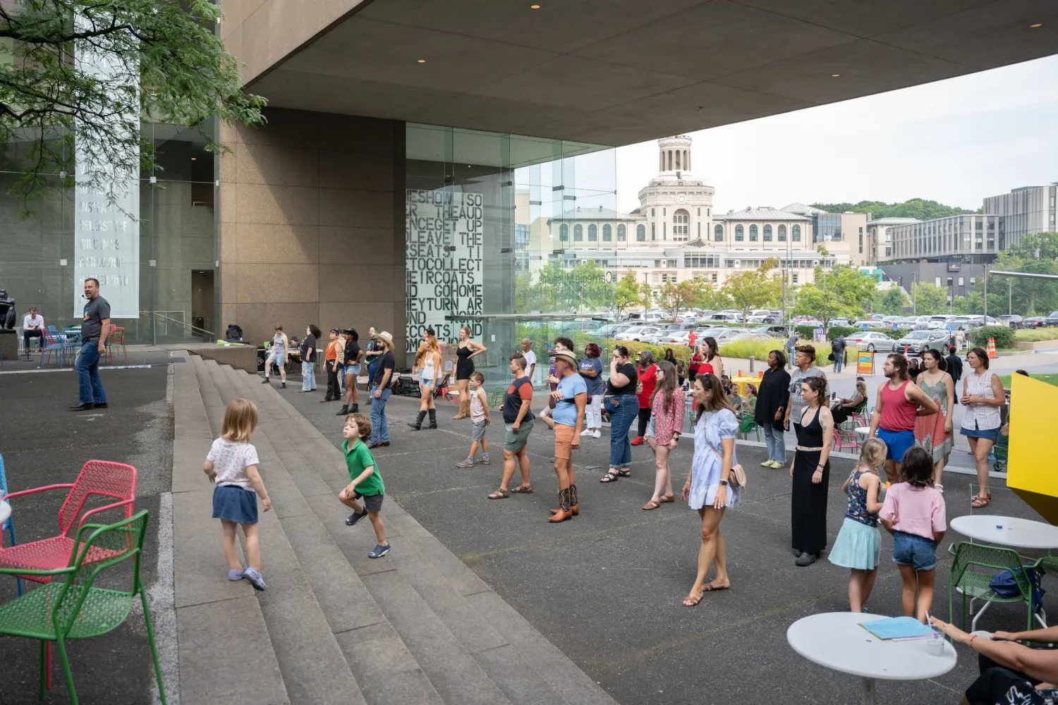 Crowd of people gathering under a covered outdoor area with green chairs and tables near a modern building.