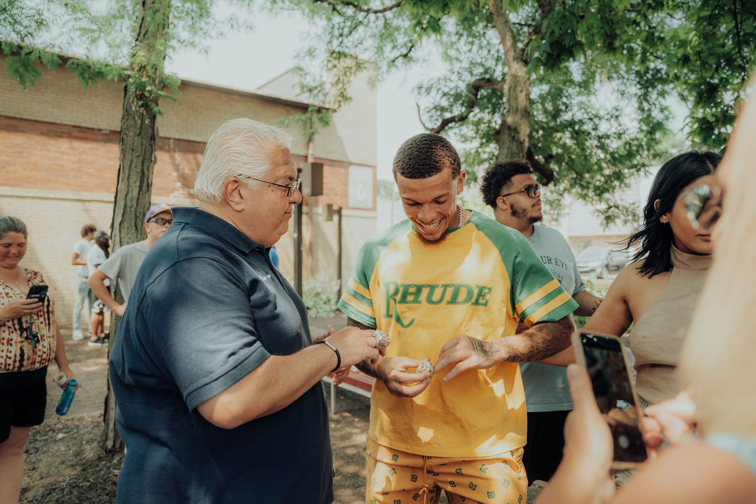 Two people interact over paperwork outdoors at a casual community gathering with others visible in the background.