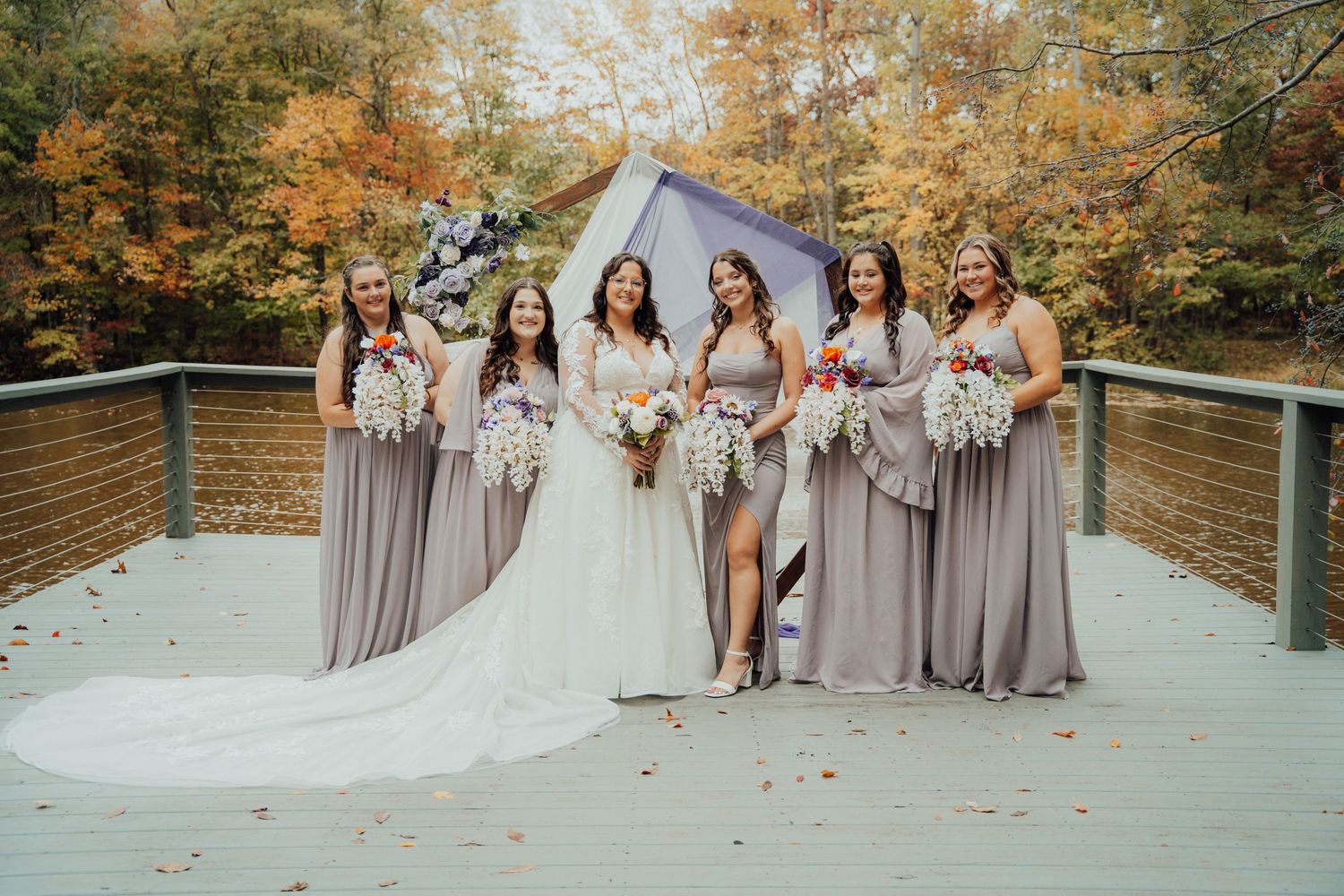 Bridal party in gray bridesmaid dresses poses on wooden bridge during autumn wedding photoshoot.