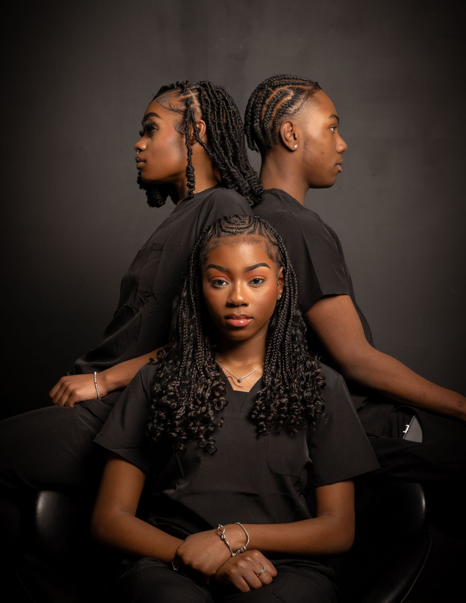 Artistic portrait of three people with braided hair wearing black poses against dark background in dramatic lighting.