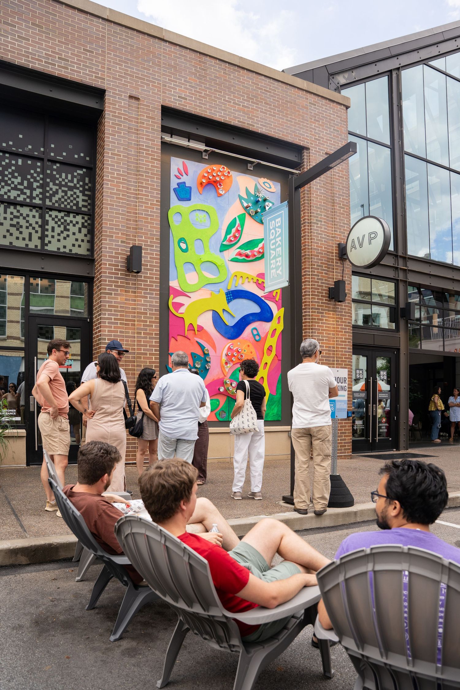 Colorful mural on brick wall outside shopping center with people gathered to view street art installation.