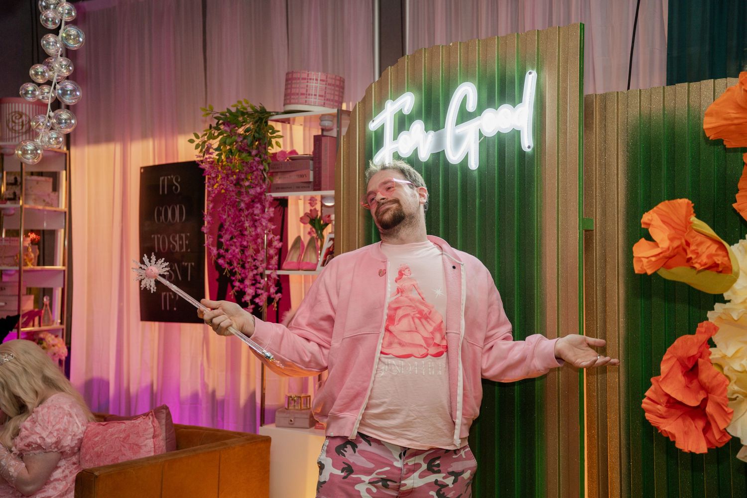 Person in pink attire gesturing with arms spread wide in front of neon signage and floral decor.