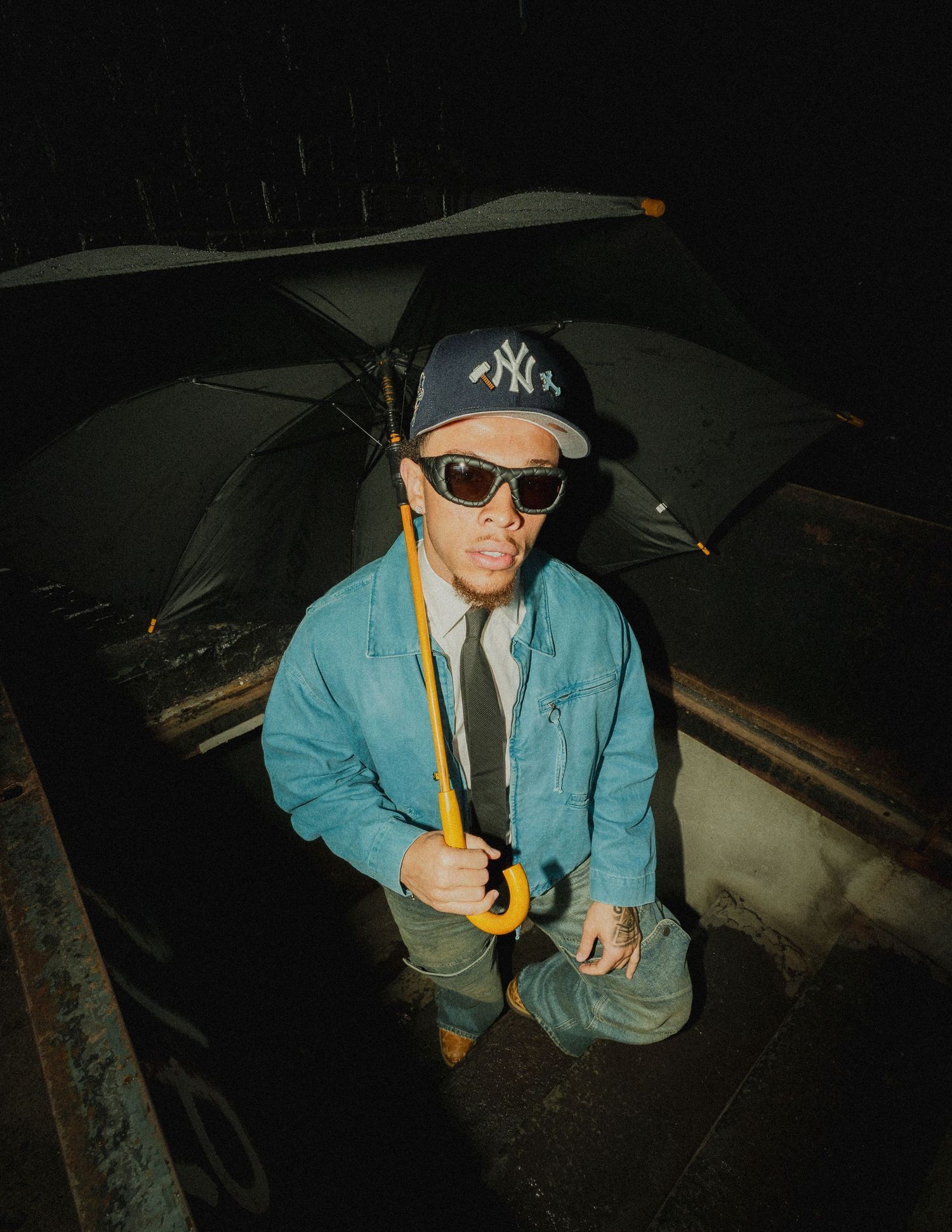 Person in a teal jacket and baseball cap crouches in a dark setting while holding a flashlight.