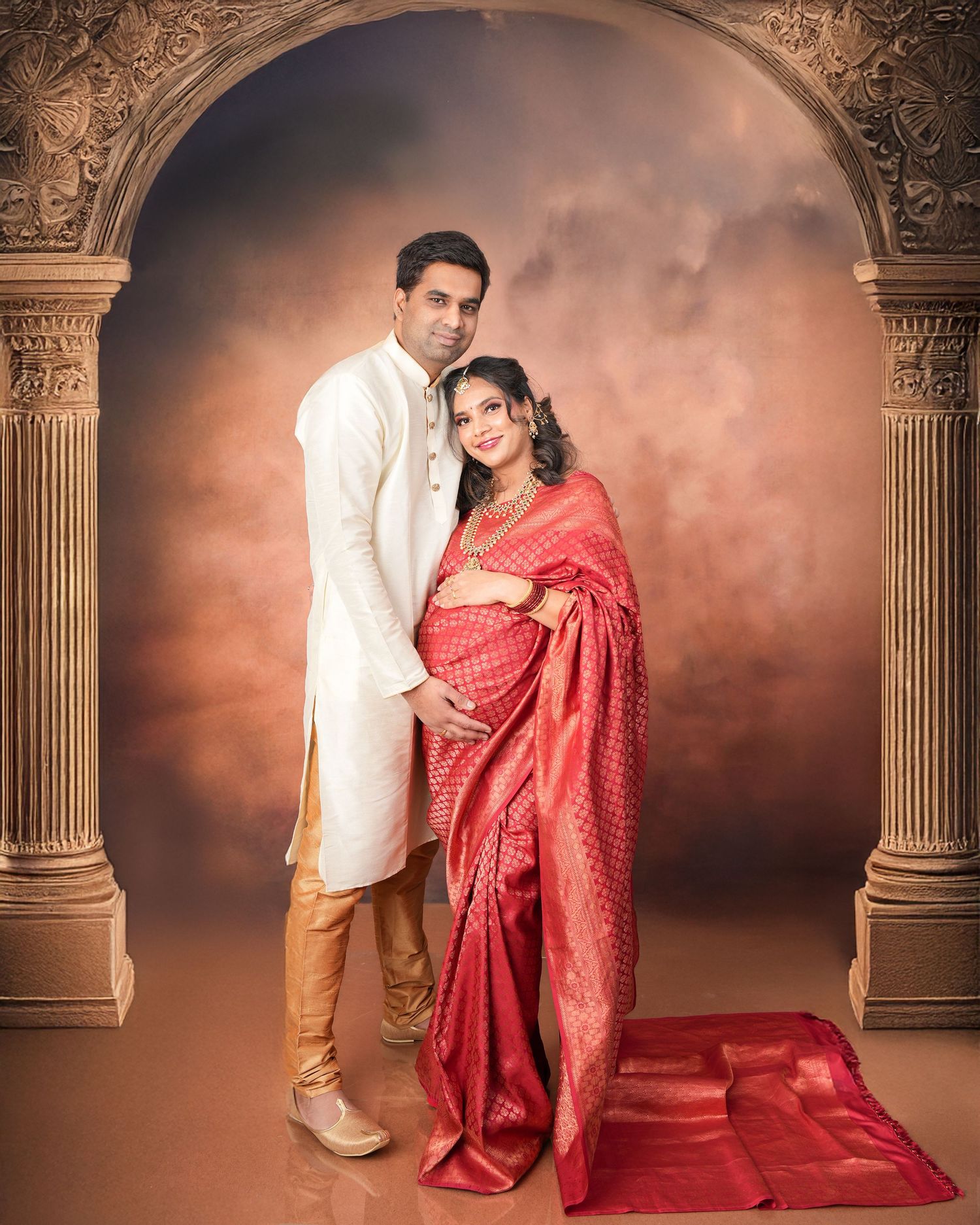 A couple in traditional Indian attire poses for a maternity photo under an ornate arch with warm lighting and dramatic backdrop.
