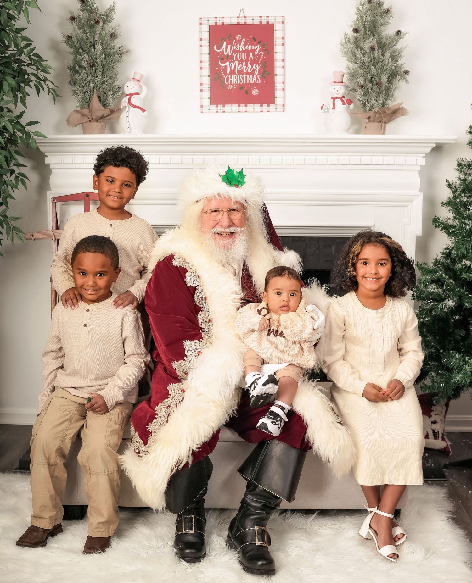 Holiday photo with Santa Claus in traditional red suit sitting with four people in cream-colored outfits by a decorated fireplace.