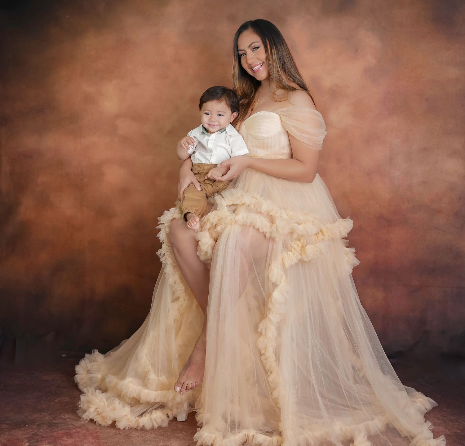 Dreamy portrait of mother and child in matching ethereal cream-colored tulle gowns against moody brown background.