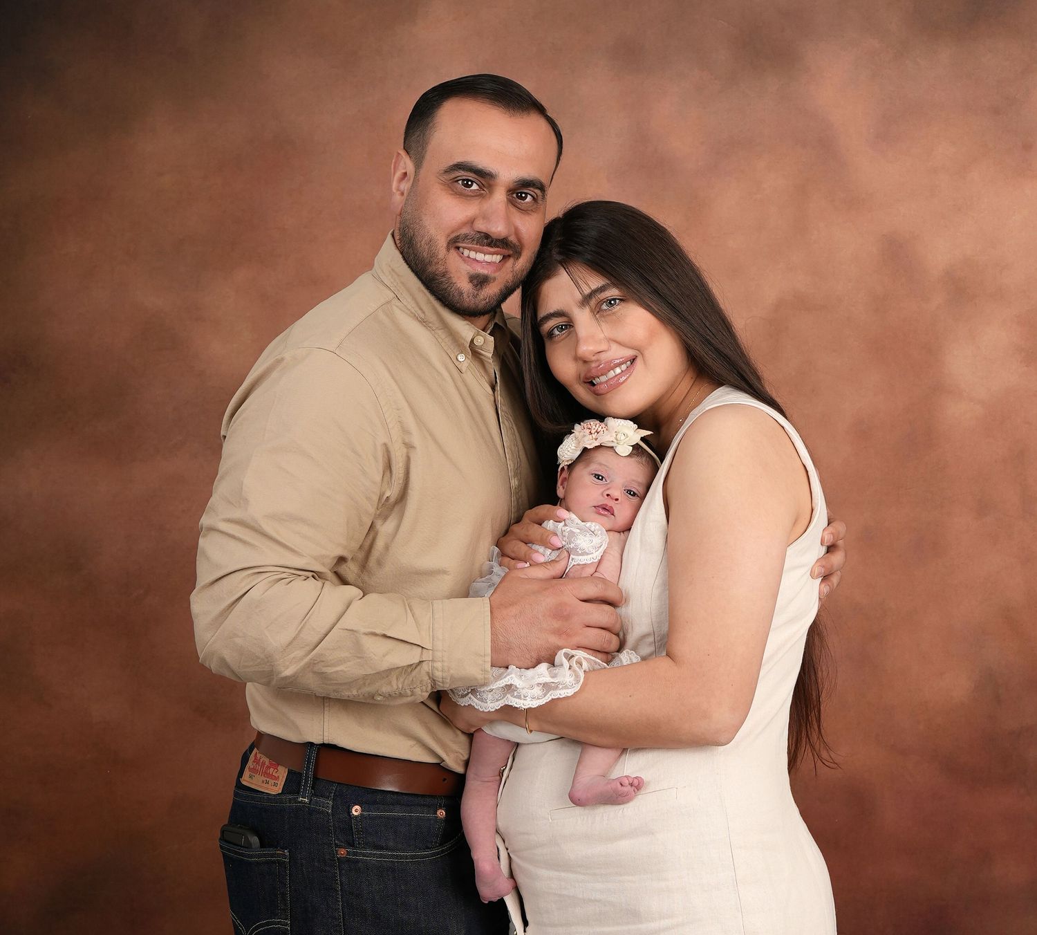 A couple in casual attire poses for a formal studio portrait while holding their newborn baby against a brown backdrop.