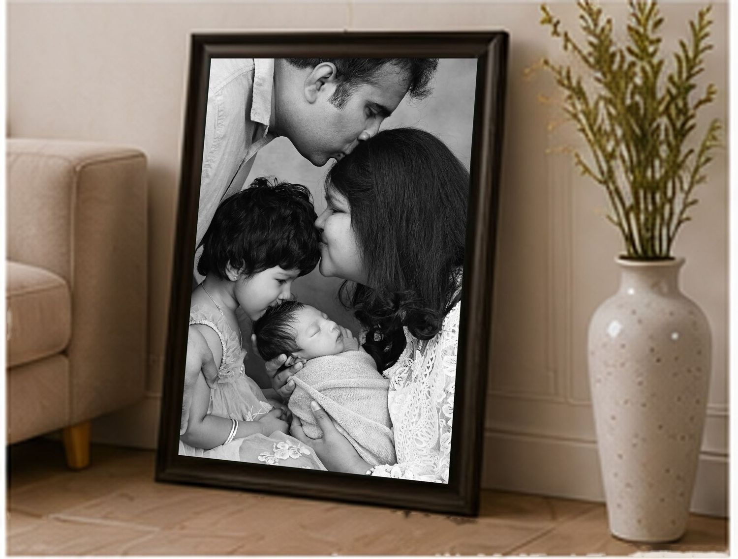 Black and white framed family photo of parents kissing newborn baby with young child, displayed on floor against wall.