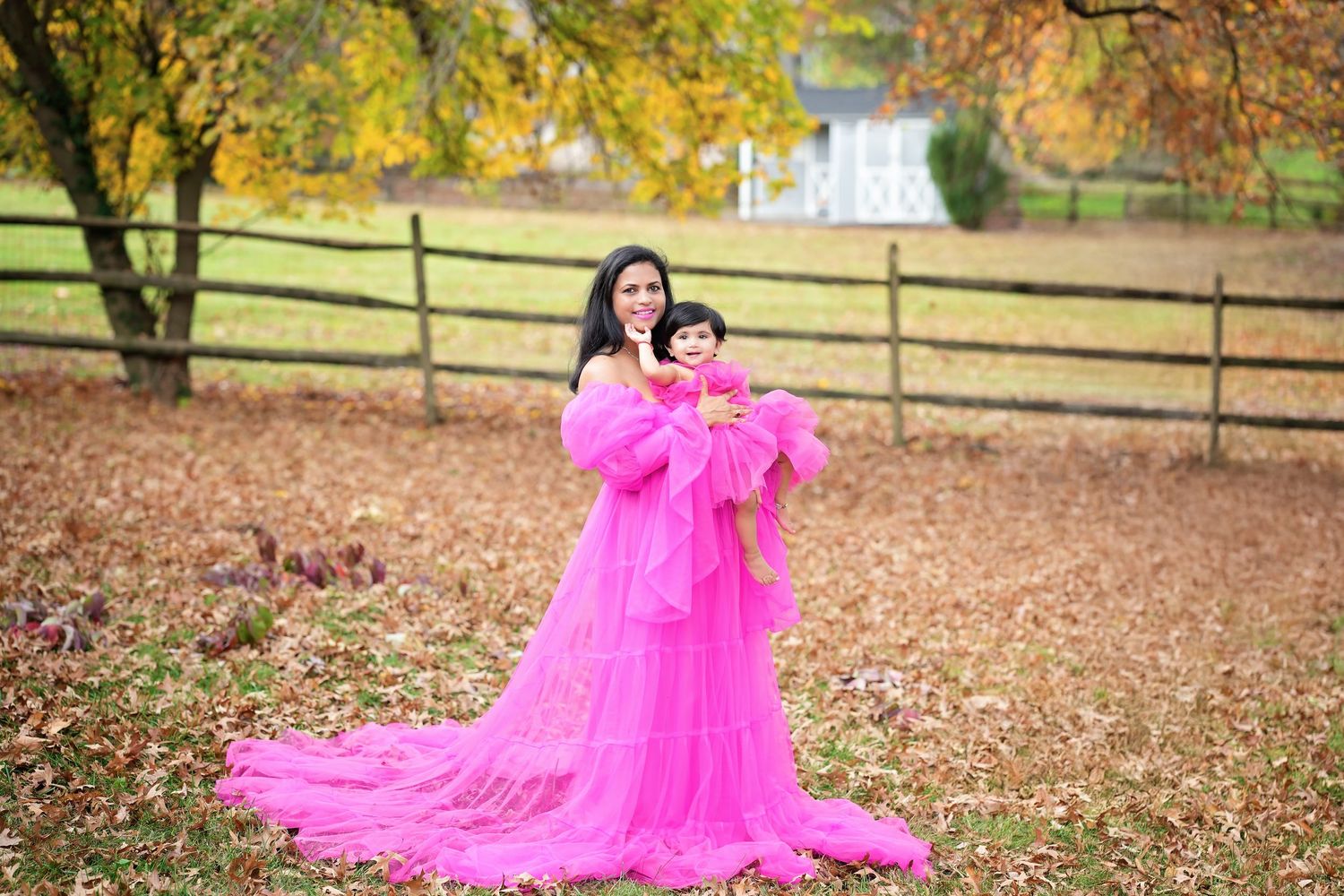Two people wearing matching bright pink tulle dresses pose in an autumn field with wooden fence and fallen leaves.