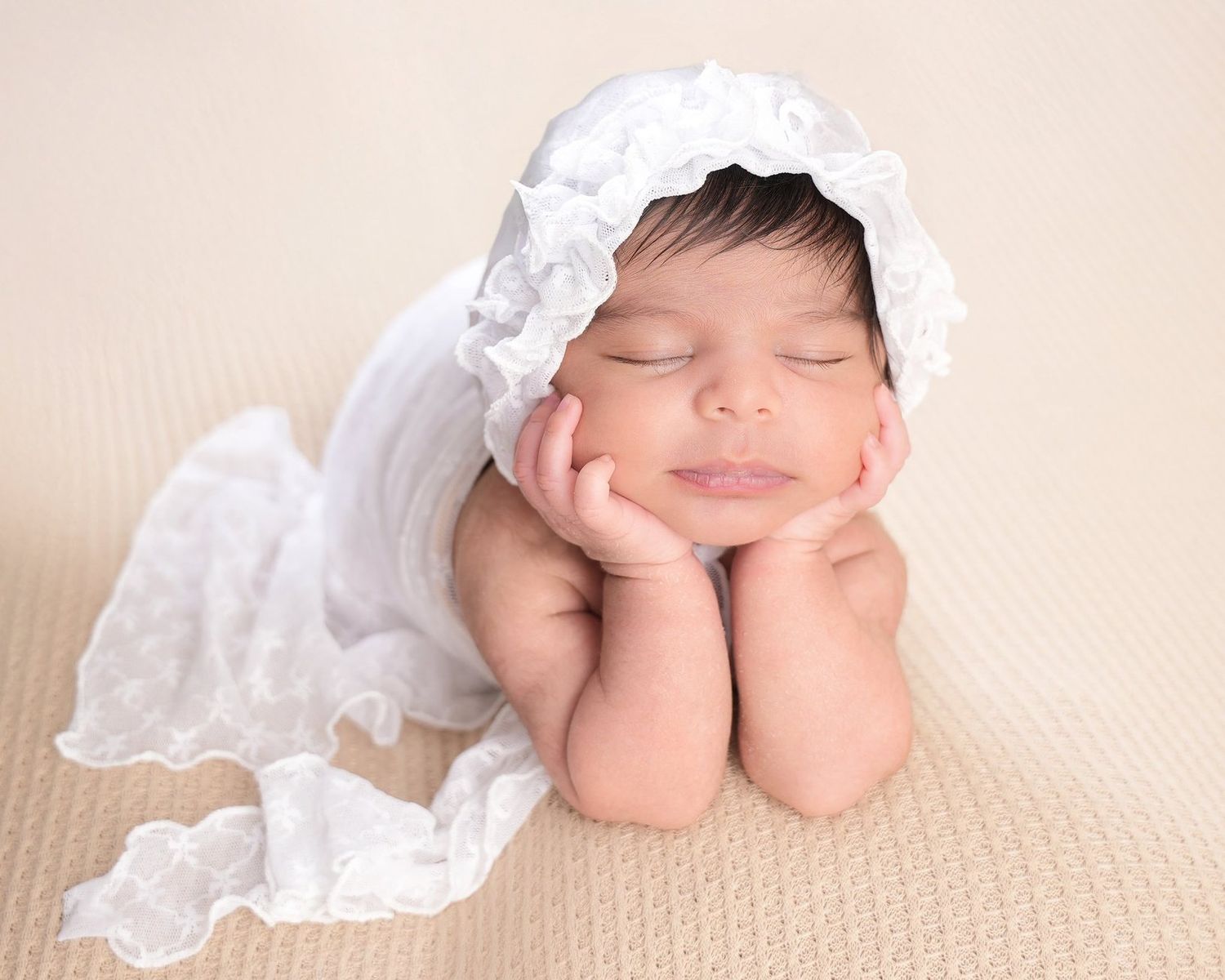 Sleeping newborn wearing white lace bonnet rests head on hands on cream textured background.