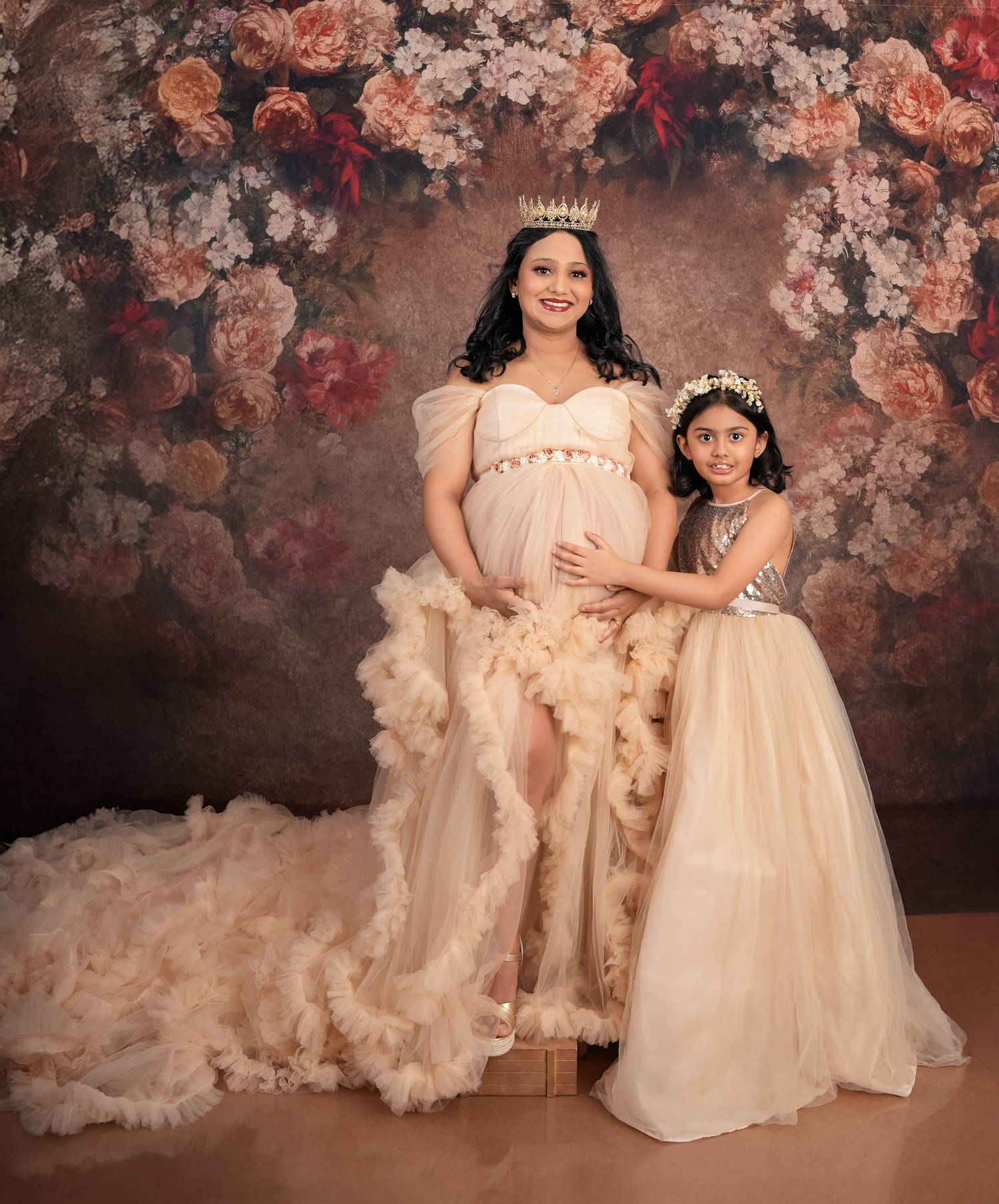 Two people in matching cream-colored formal dresses pose together against a moody floral backdrop.
