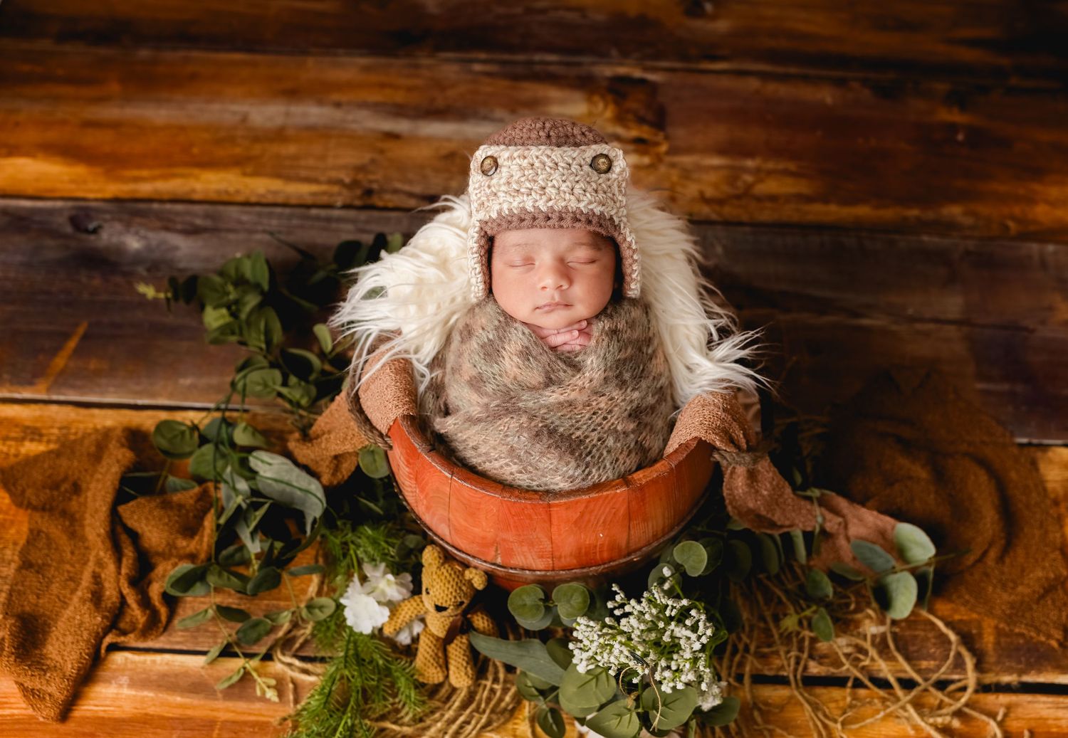 A newborn in a knit hat rests in an orange bowl surrounded by greenery and flowers on a rustic wooden floor.