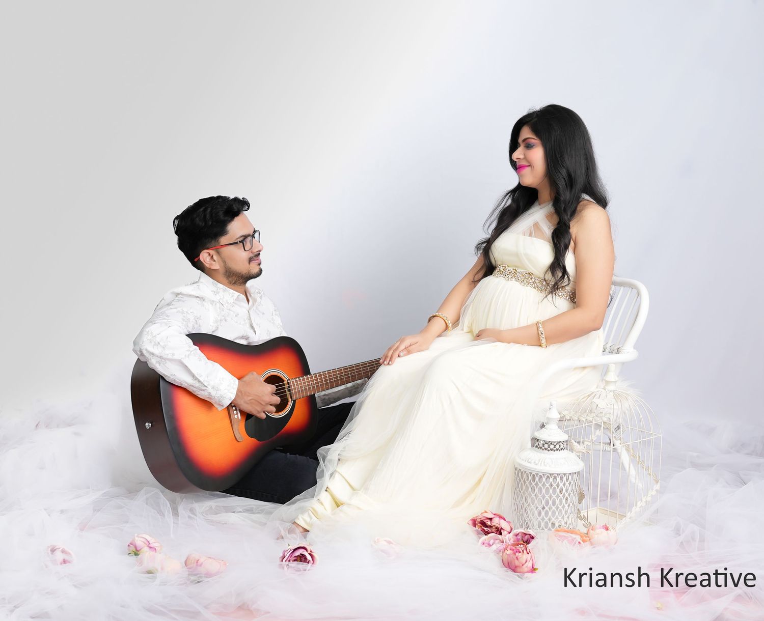 A couple in white outfits sits together during a romantic photoshoot with a guitar and scattered rose petals.