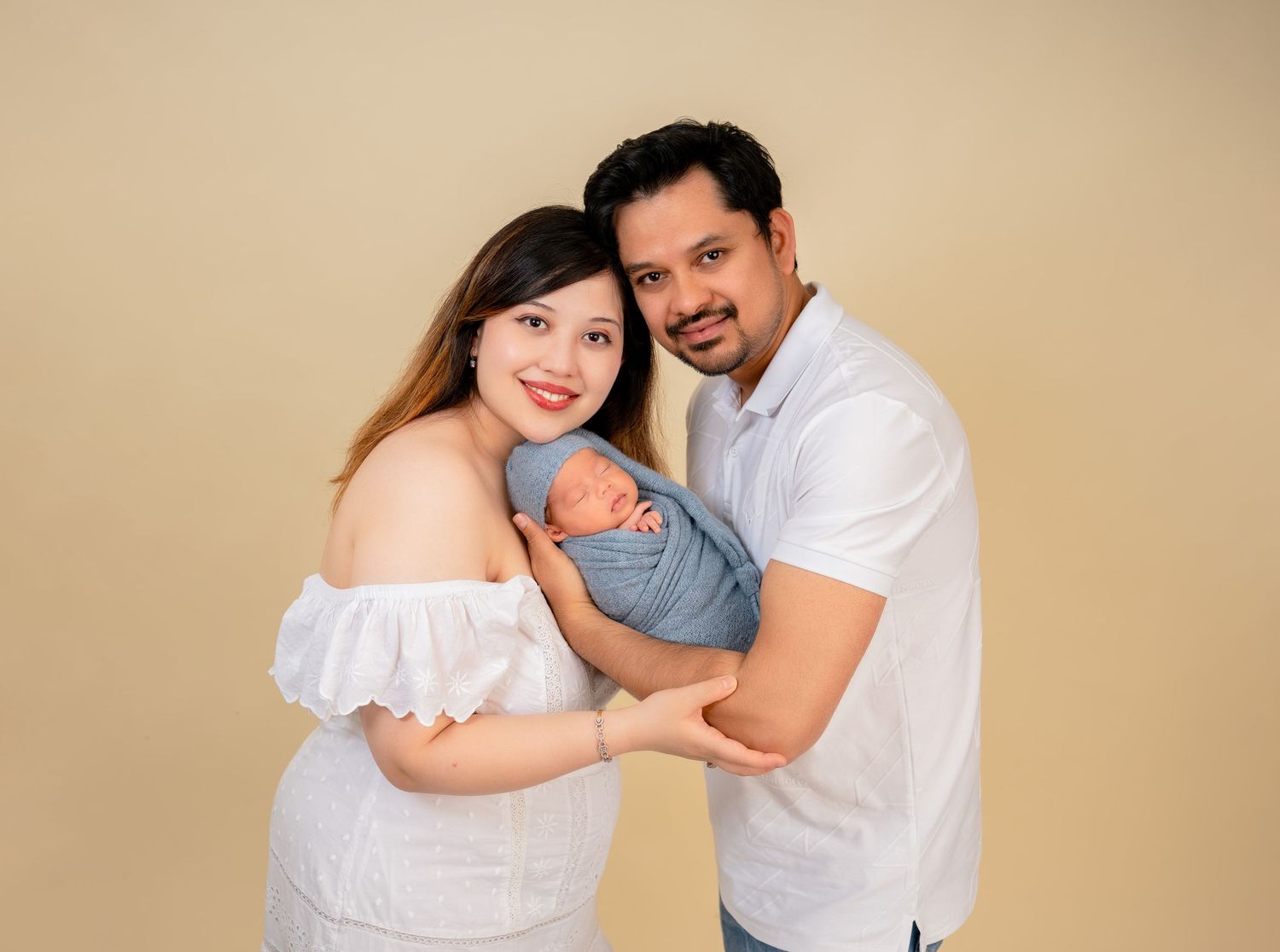 Happy couple in white outfits holding their newborn baby wrapped in blue against a beige studio background.