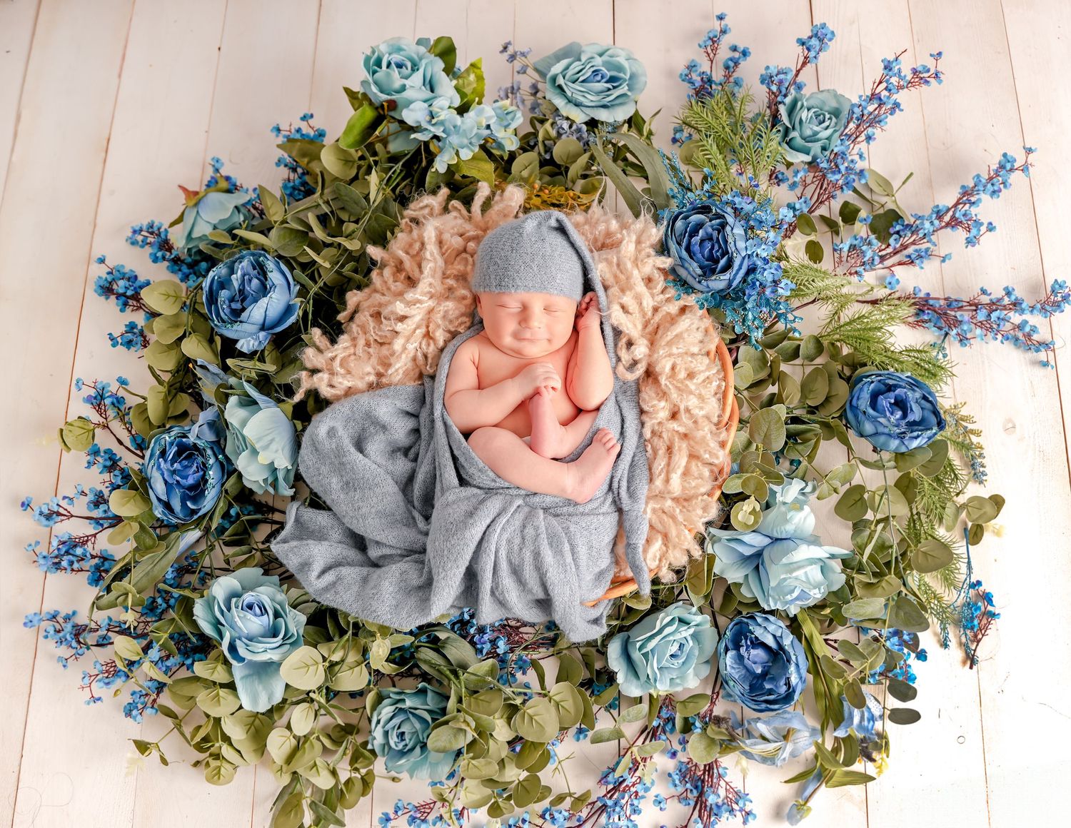 Sweet newborn photo surrounded by a floral wreath featuring blue roses and delicate greenery on wooden floorboards.