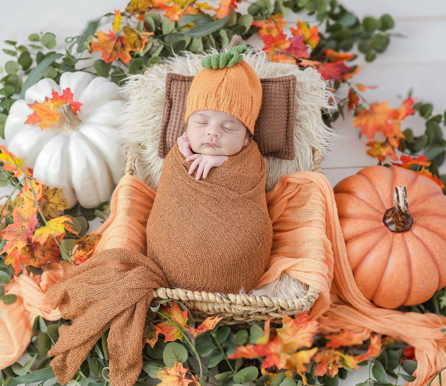Cozy fall newborn photoshoot setup with white and orange pumpkins, autumn leaves, and rustic wooden elements.