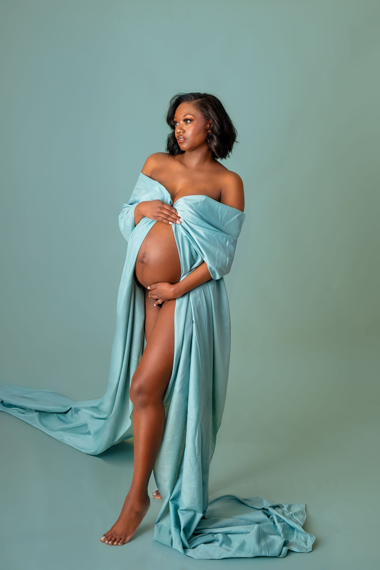 Artistic maternity photo series with flowing light blue fabric in minimalist studio setting against sage green backdrop.