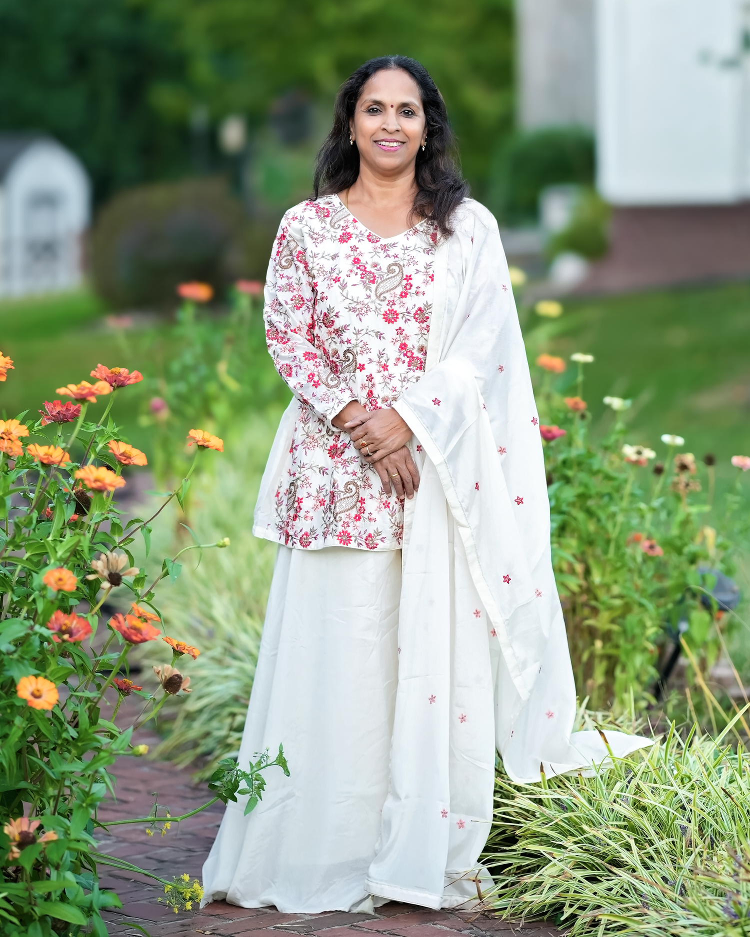 Person wearing a white and red floral dress with matching white dupatta standing in a garden with orange flowers.