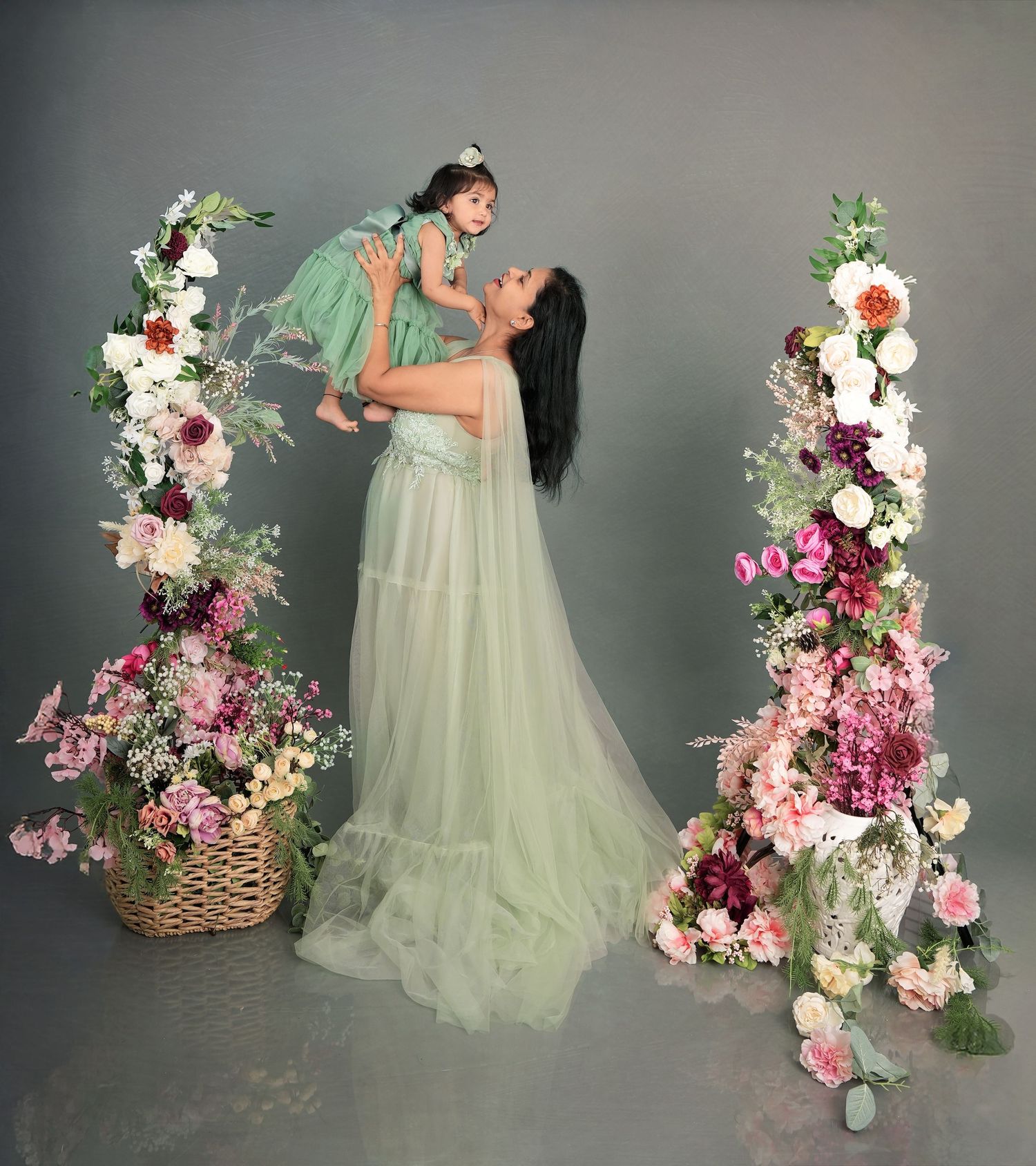 A figure in a sage green tulle dress poses elegantly with pink and white floral arrangements against a gray backdrop.
