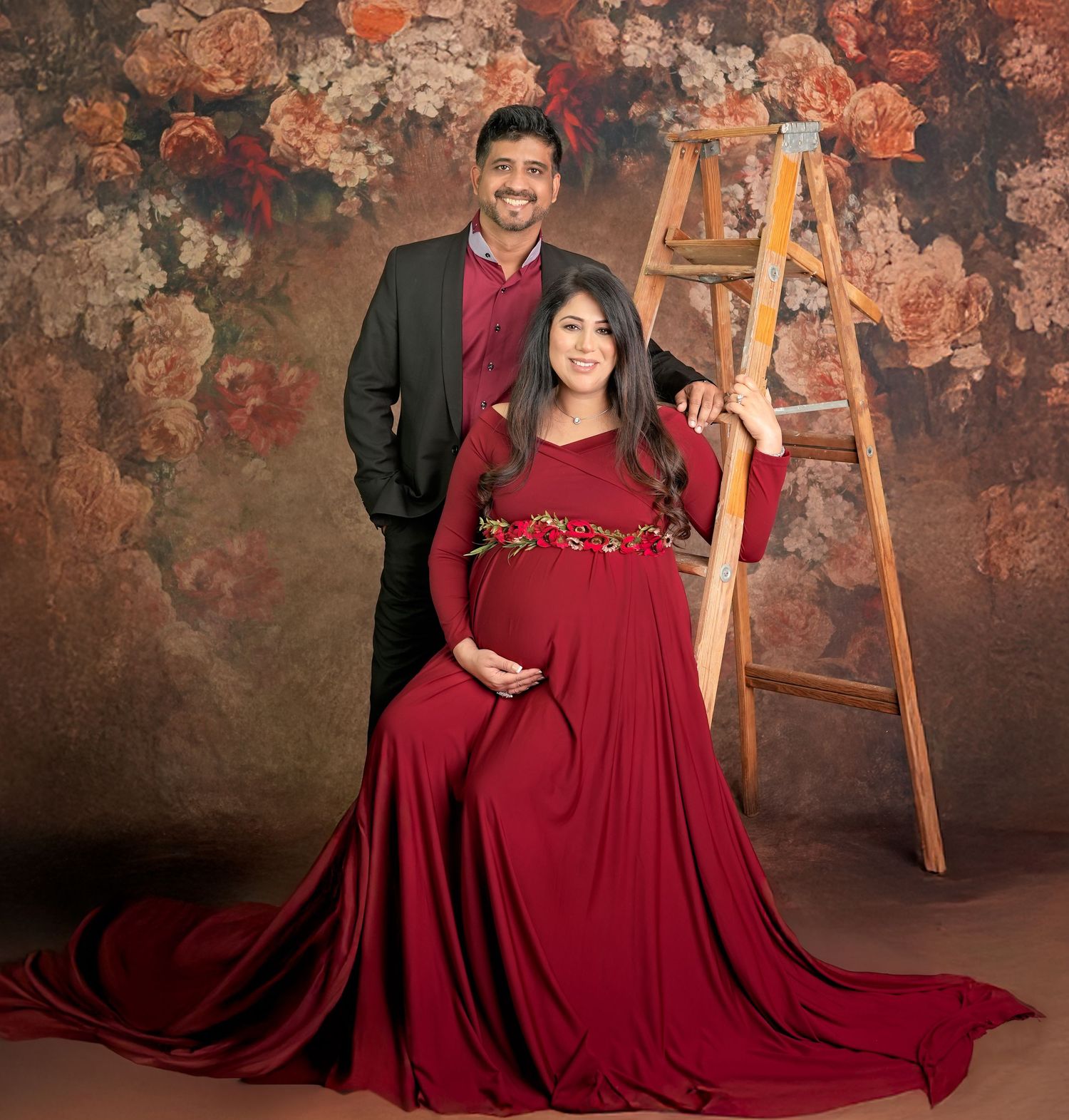 A couple poses in burgundy formal attire against a textured dark backdrop with an antique gold easel.