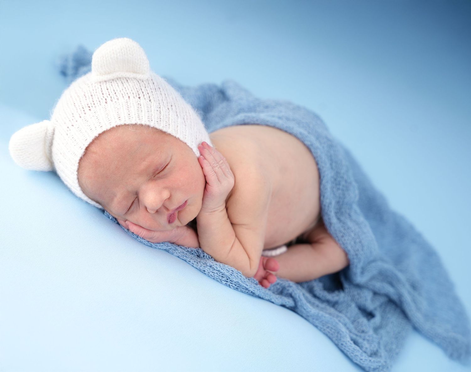 A sleeping newborn baby wrapped in soft blue blankets wearing a white knit hat rests peacefully.