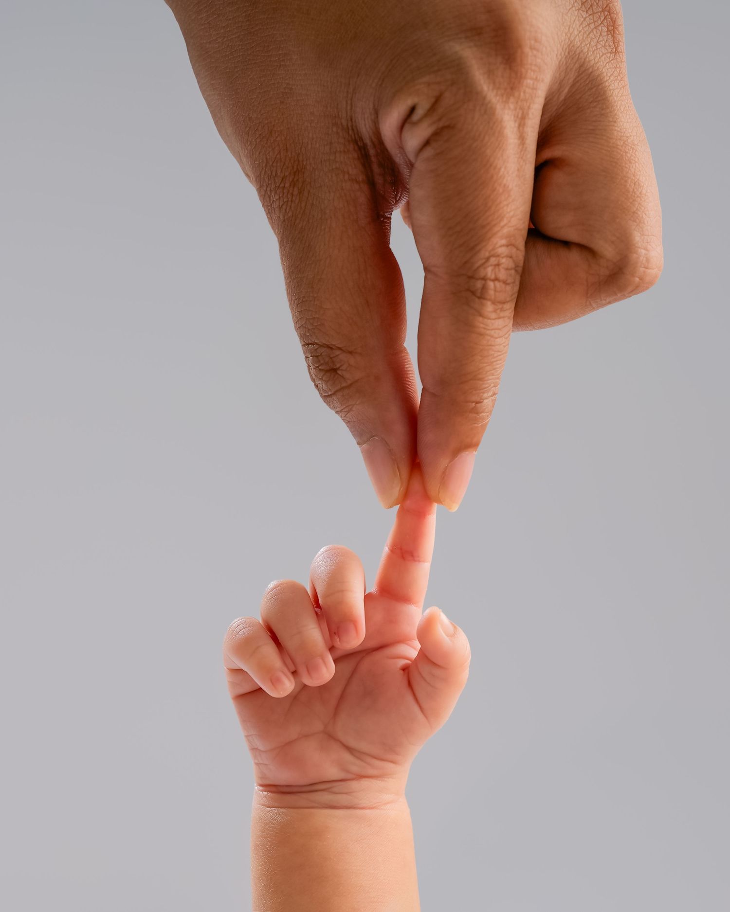 Adult's hand touching a baby's tiny outstretched finger in a tender gesture against a gray background.