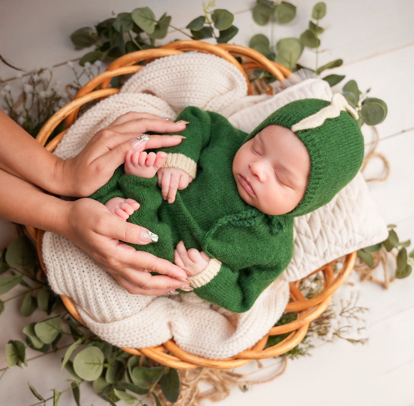 Newborn wrapped in cream knit blanket rests in wicker basket surrounded by delicate eucalyptus leaves and greenery.